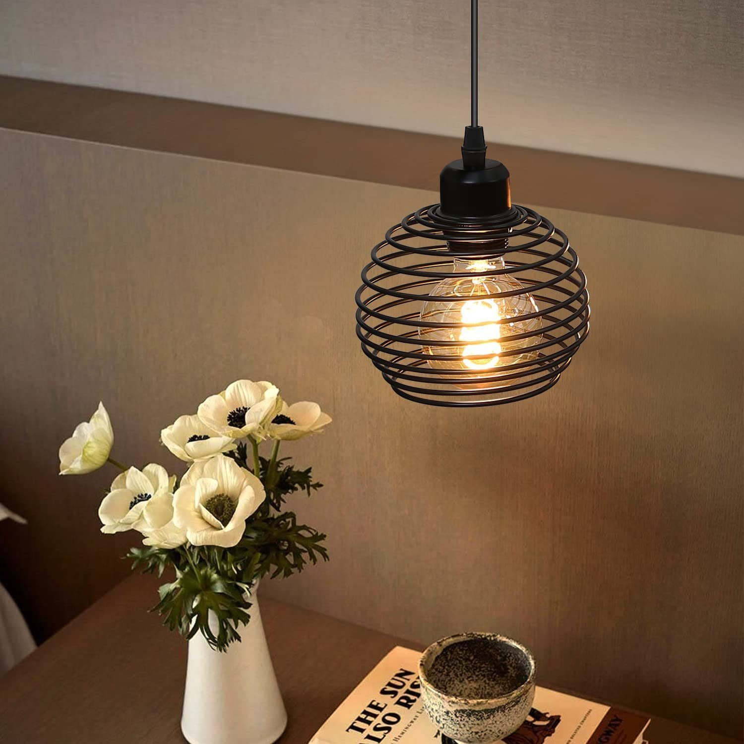 Czarna metalowa lampa wisząca z drucianą ramą z widoczną żarówką, nad kwiatami i książką.