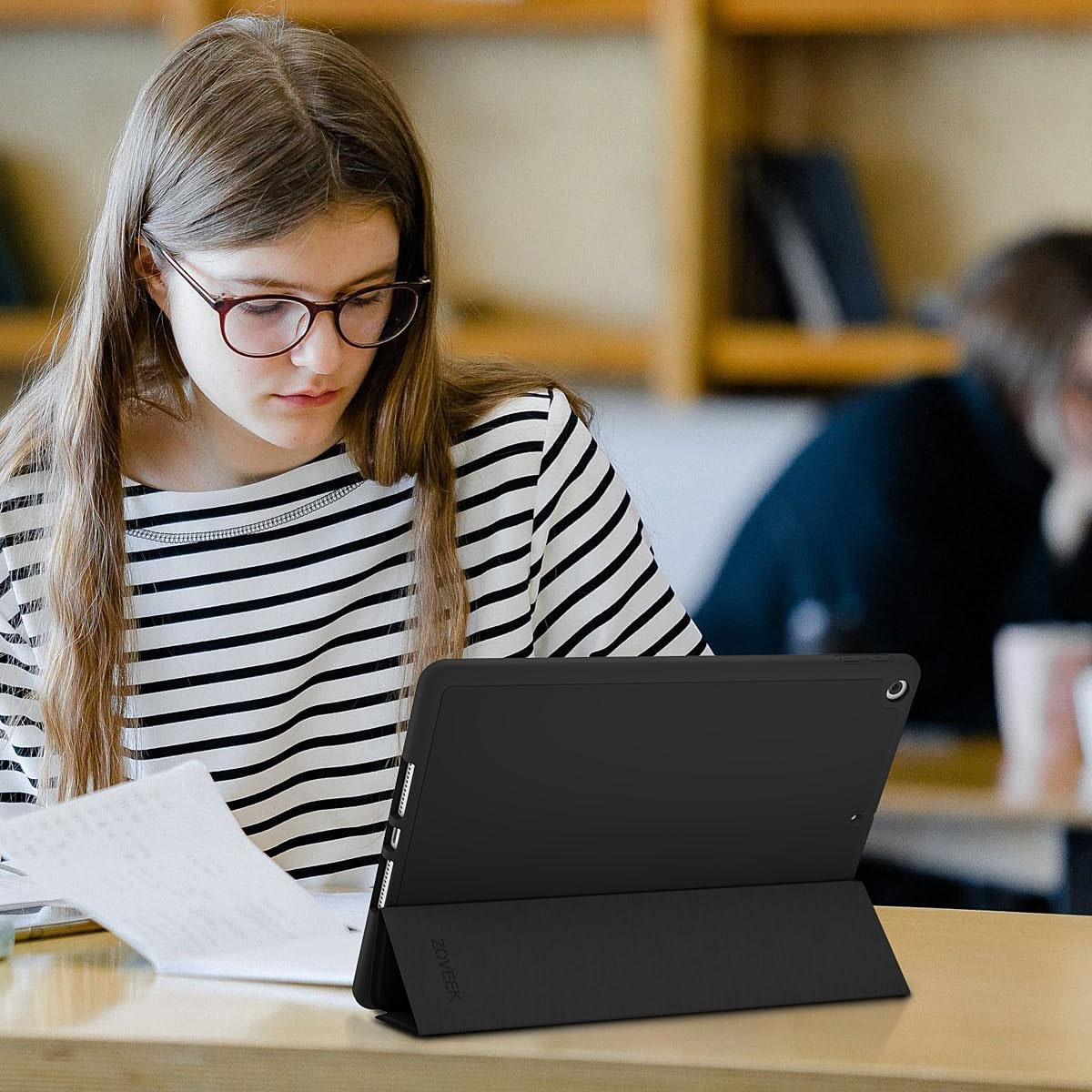 Eine junge Frau mit Brille, die auf ein schwarzes Tablet blickt. Ein Bibliotheks-Hintergrund mit einer weiteren Person unscharf.