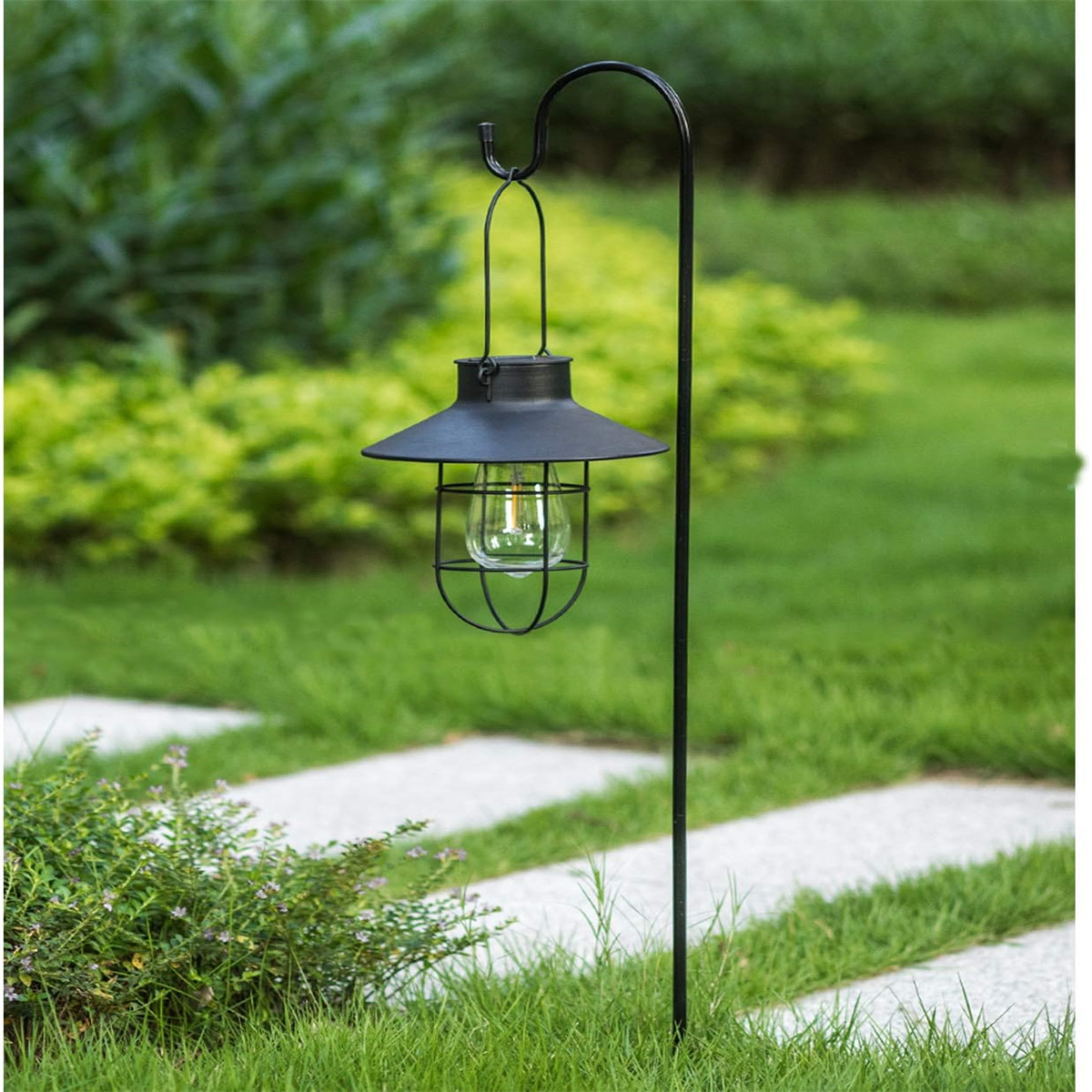 Schwarze Außenlaterne mit gebogenem Metallpfahl. Grünes Gras und Büsche im Hintergrund.