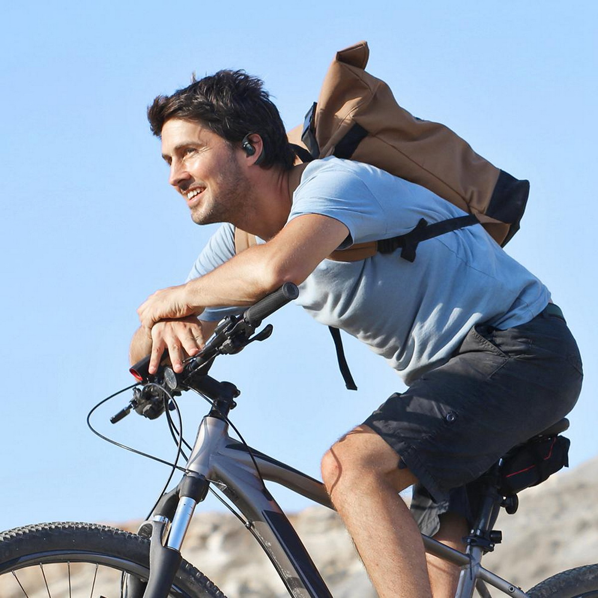 Mann fährt Mountainbike auf einem Trail. Er trägt ein blaues Hemd und einen Rucksack. Der Himmel ist klar.