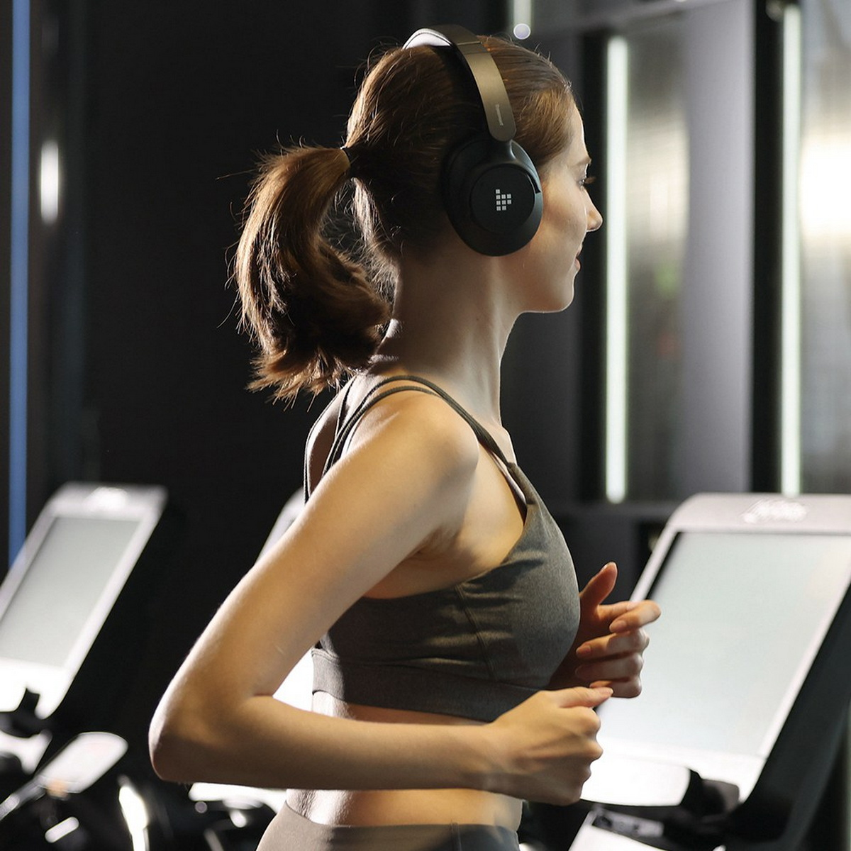 Frau im Fitnessstudio läuft auf einem Laufband und trägt Kopfhörer. Andere Laufbänder im Hintergrund.