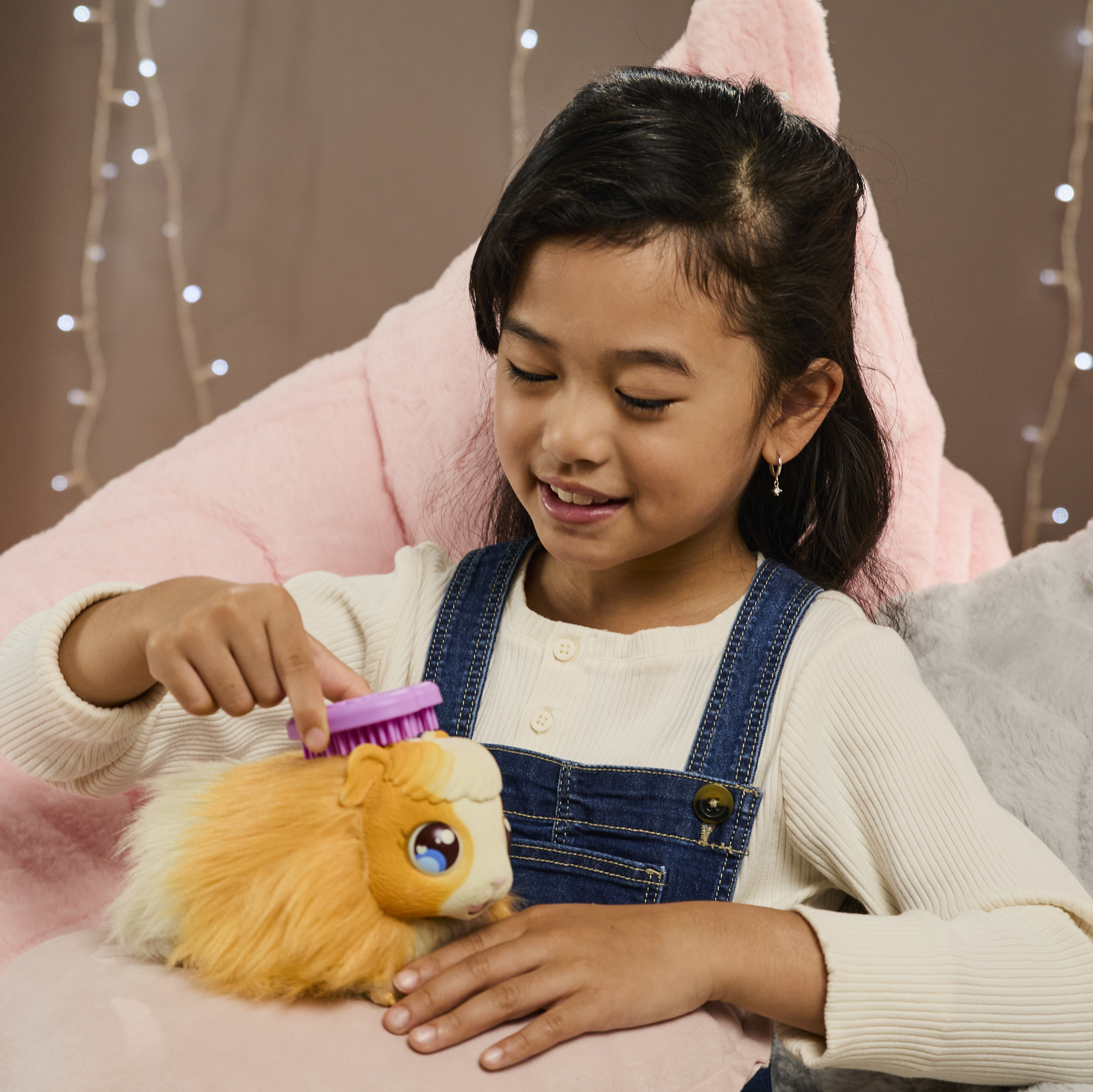 Dziewczynka szczotkuje zabawkę świnkę morską. Zabawka jest pomarańczowa i biała. Różowe puszyste tło.