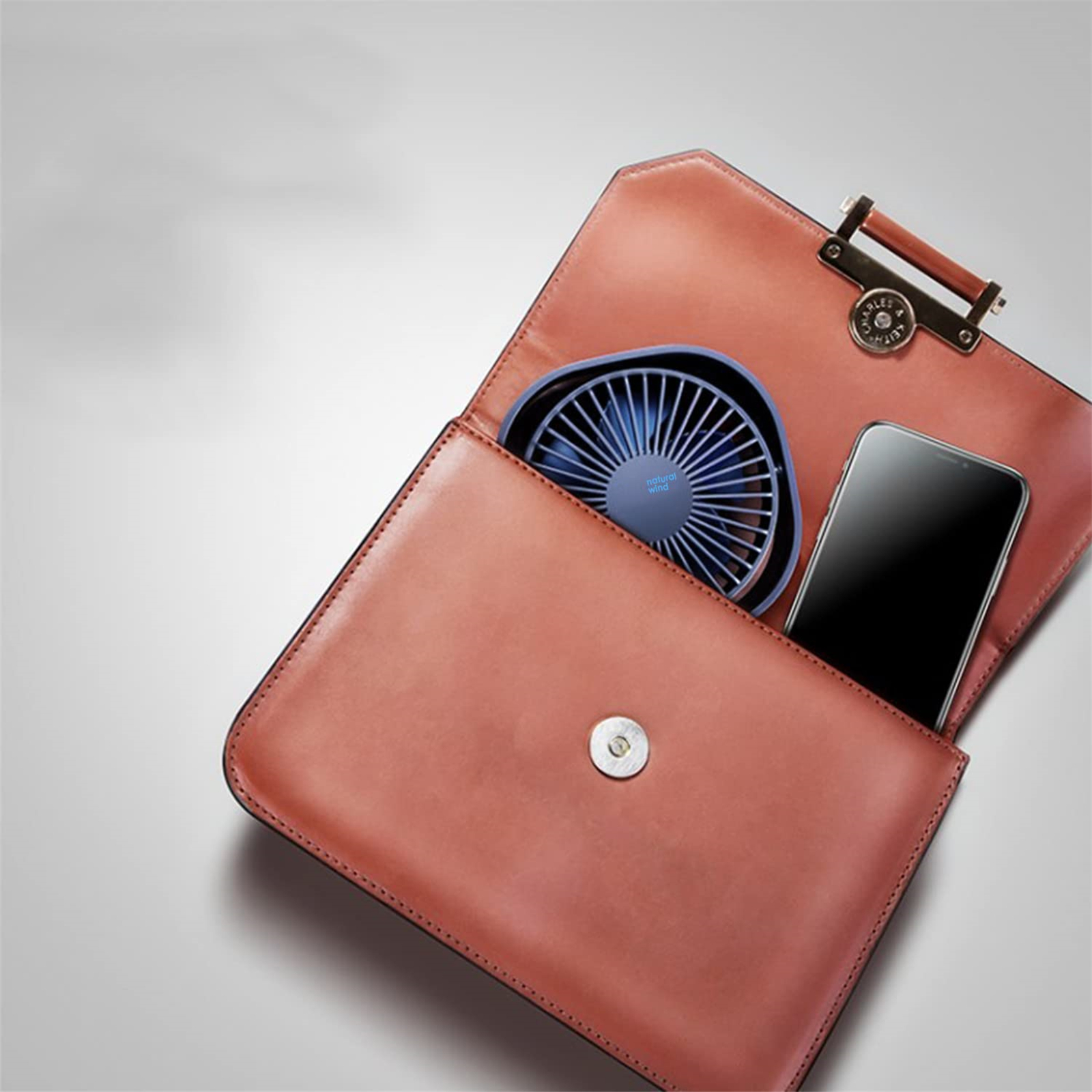 Ein kleiner blauer Ventilator und ein Smartphone liegen in einer Handtasche. Die Handtasche ist braun.