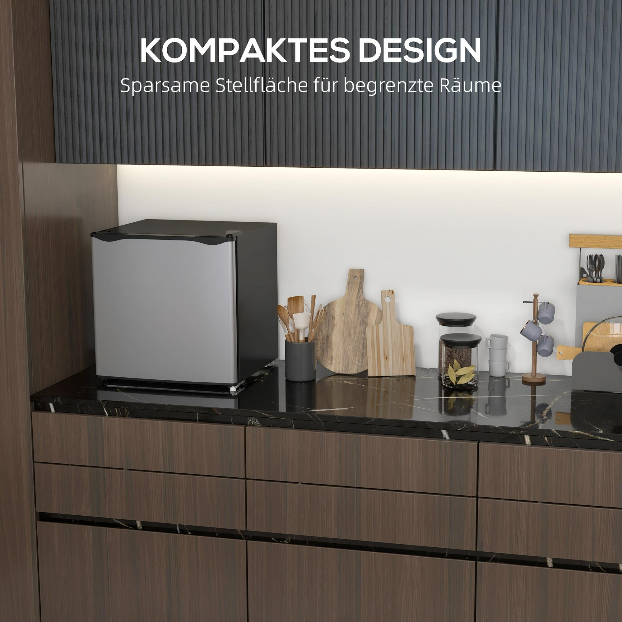 Küchenszene mit einem kleinen silbernen Kühlschrank. Weitere Gegenstände sind Schneidebretter, Gläser und Utensilien.