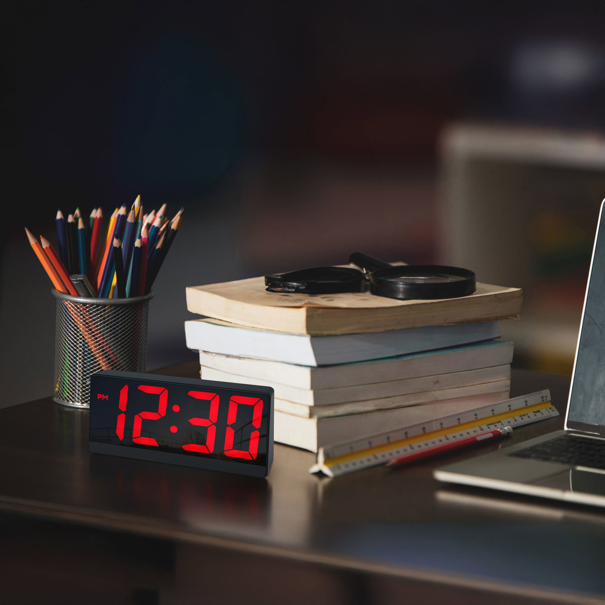 Digitaler Wecker, zeigt 12:30 PM in roten Ziffern. Auf einem Schreibtisch mit Büchern, Bleistiften und Laptop.