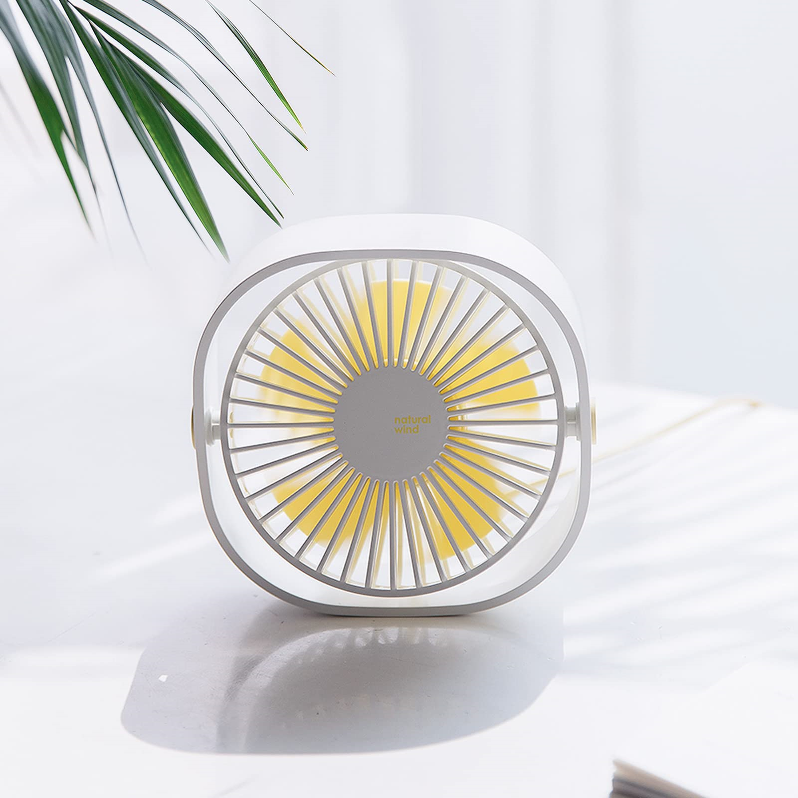 Weißer und grauer Ventilator mit gelben Flügeln auf weißer Oberfläche. Im Hintergrund grüne Blätter.