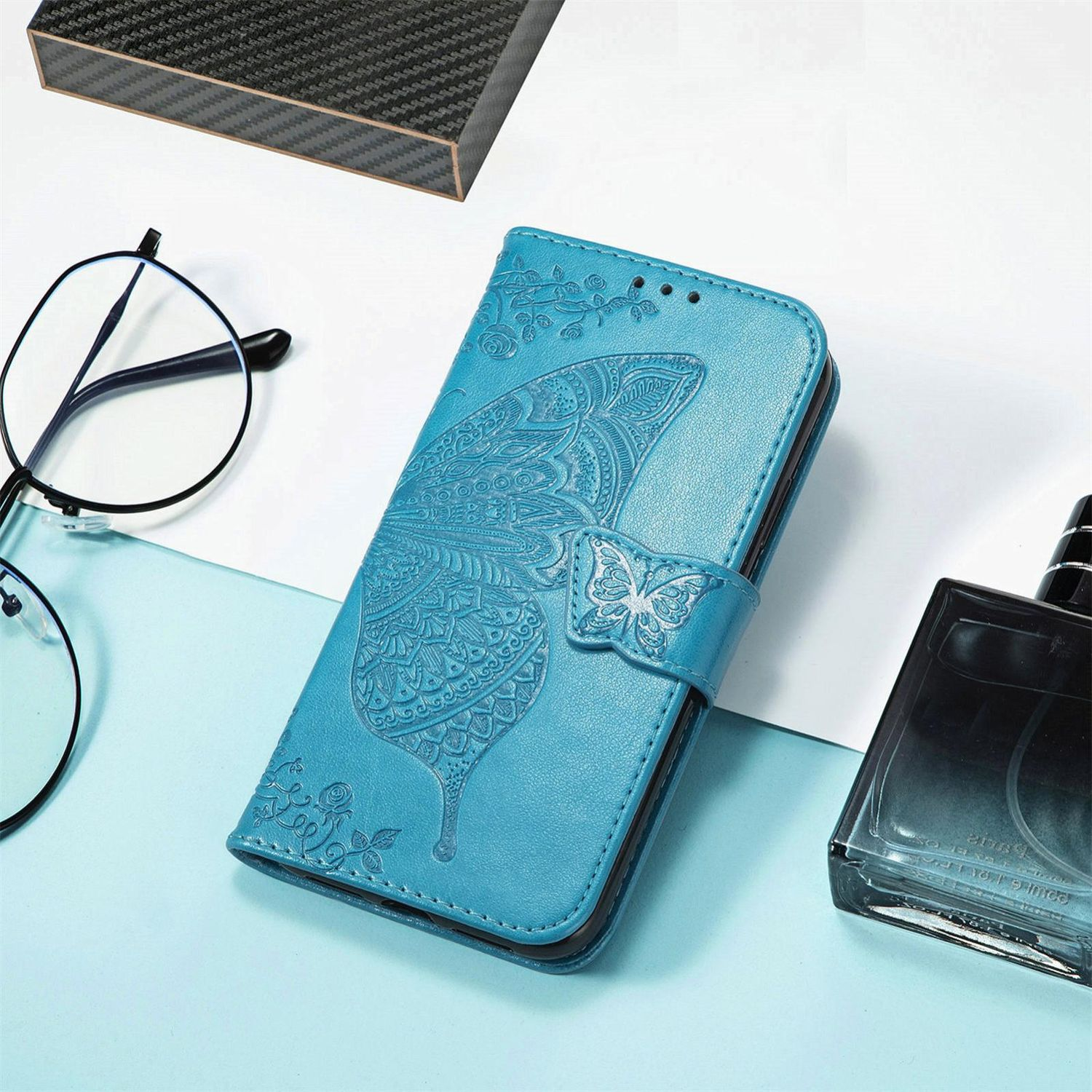 Funda azul para teléfono con diseño de mariposa y gafas. Una caja negra y un frasco de perfume también son visibles.