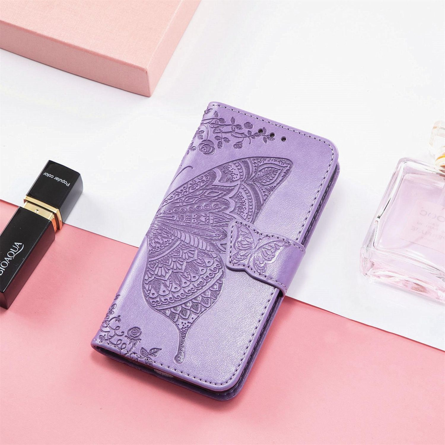 Funda morada para teléfono con diseño de mariposa, cerrada. Hay un lápiz labial y un frasco de perfume al lado.
