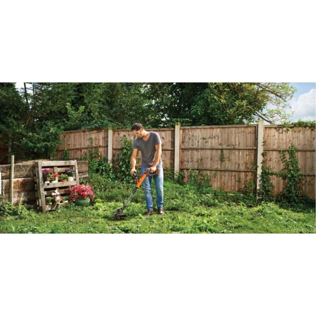 Mężczyzna używający podkaszarki w ogrodzie. Drewniane ogrodzenie i rośliny w tle.