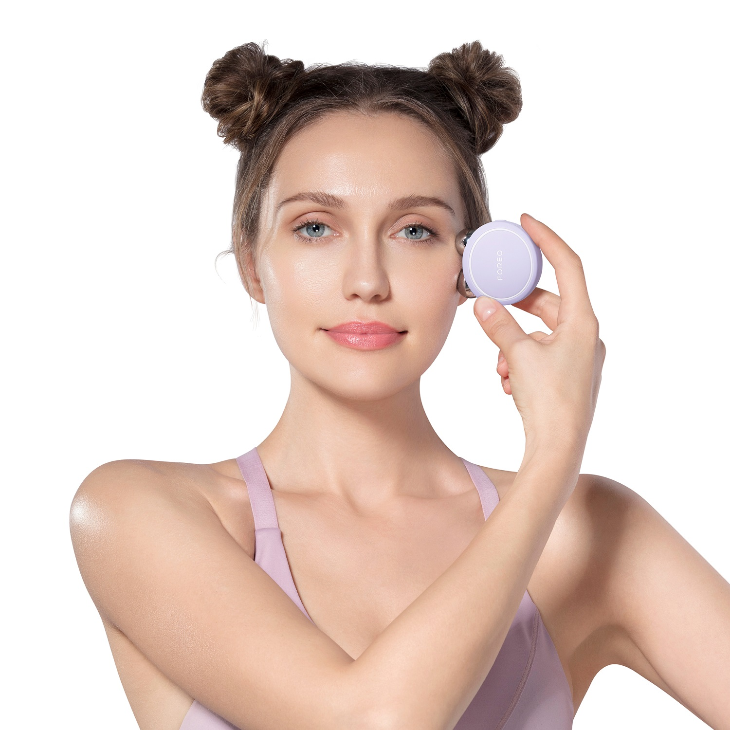 Una donna tiene in mano un massaggiatore facciale viola. Ha degli space buns e indossa un top lilla chiaro.