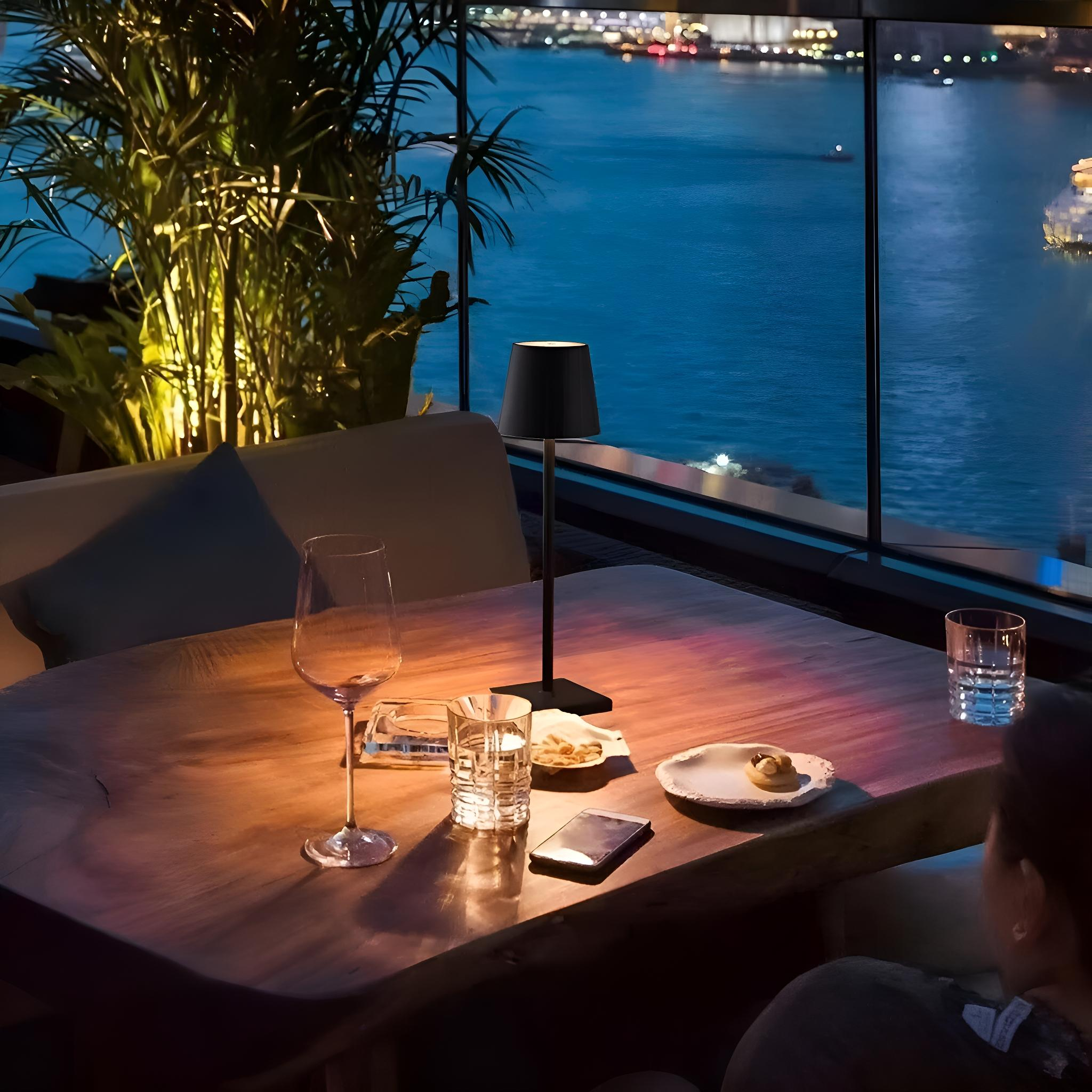 Schwarze Lampe auf einem Tisch mit Blick auf Wasser und Gebäude. Gläser und andere Gegenstände sind auch auf dem Tisch.