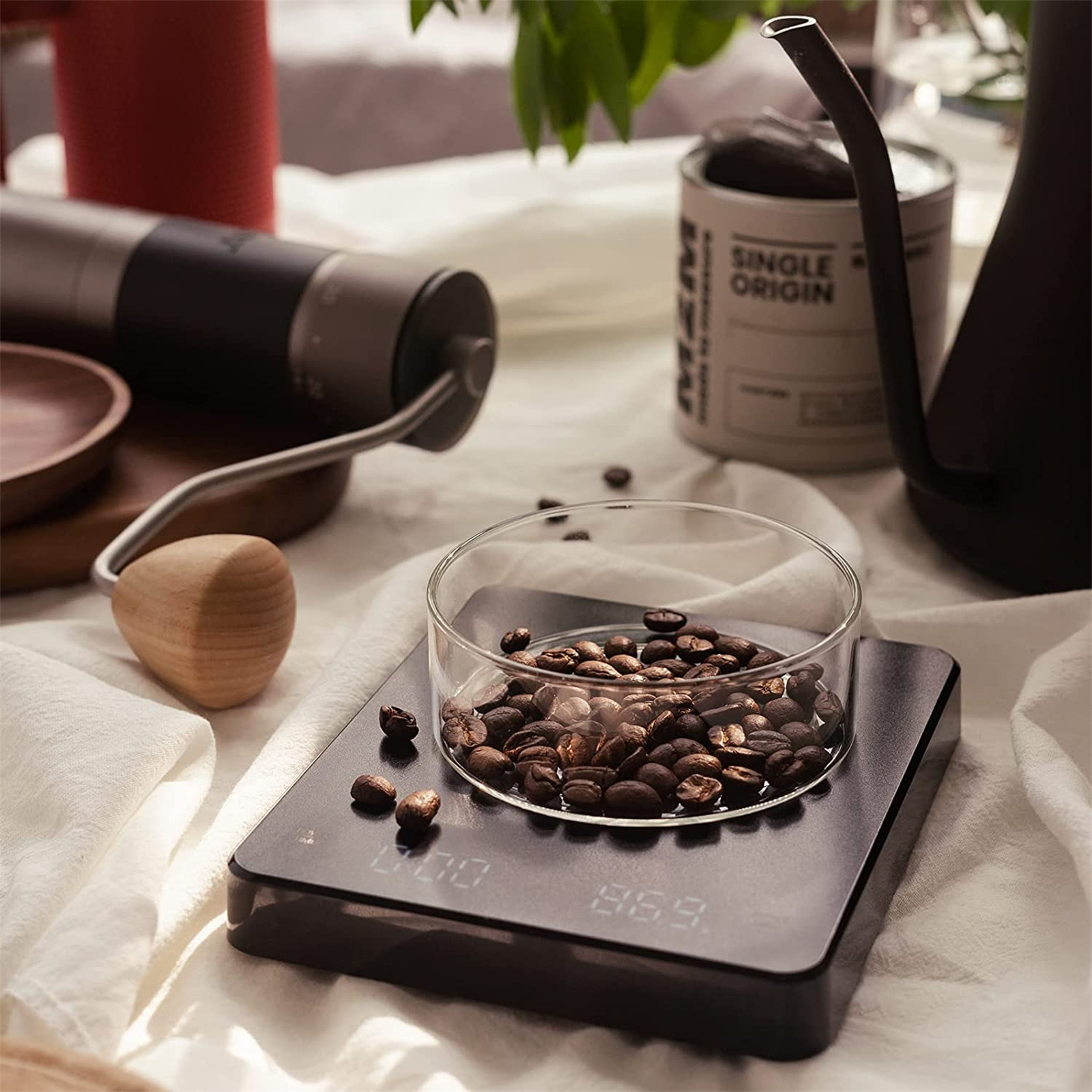 Kaffeebohnen in einer Glasschale auf einer Waage, mit einer Mühle und anderen Gegenständen im Hintergrund.