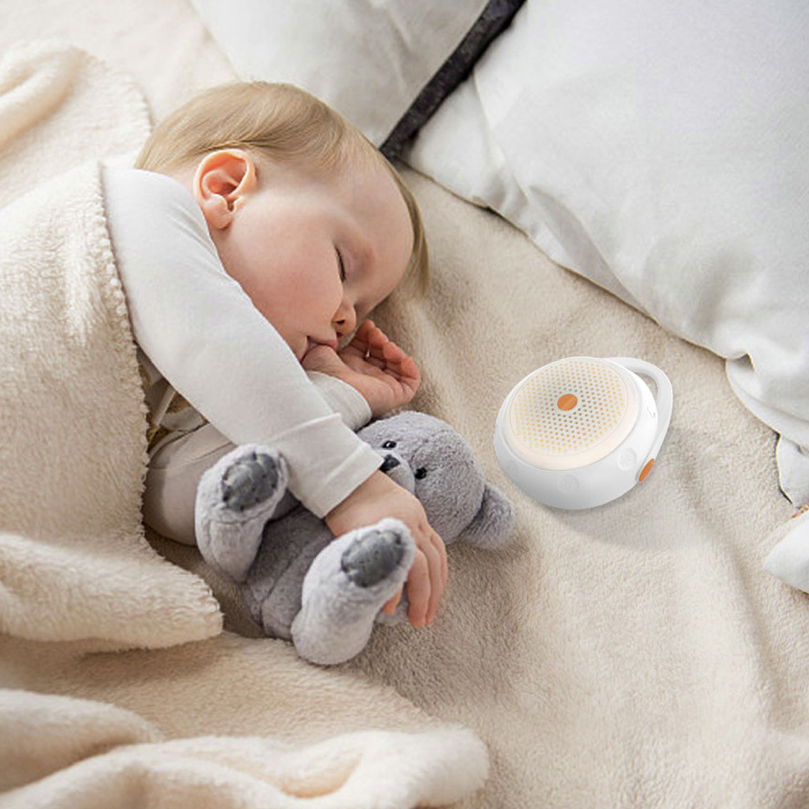 Ein Baby schläft neben einer weißen und orangefarbenen Soundmaschine auf einem Bett, mit einem grauen Teddybären daneben.