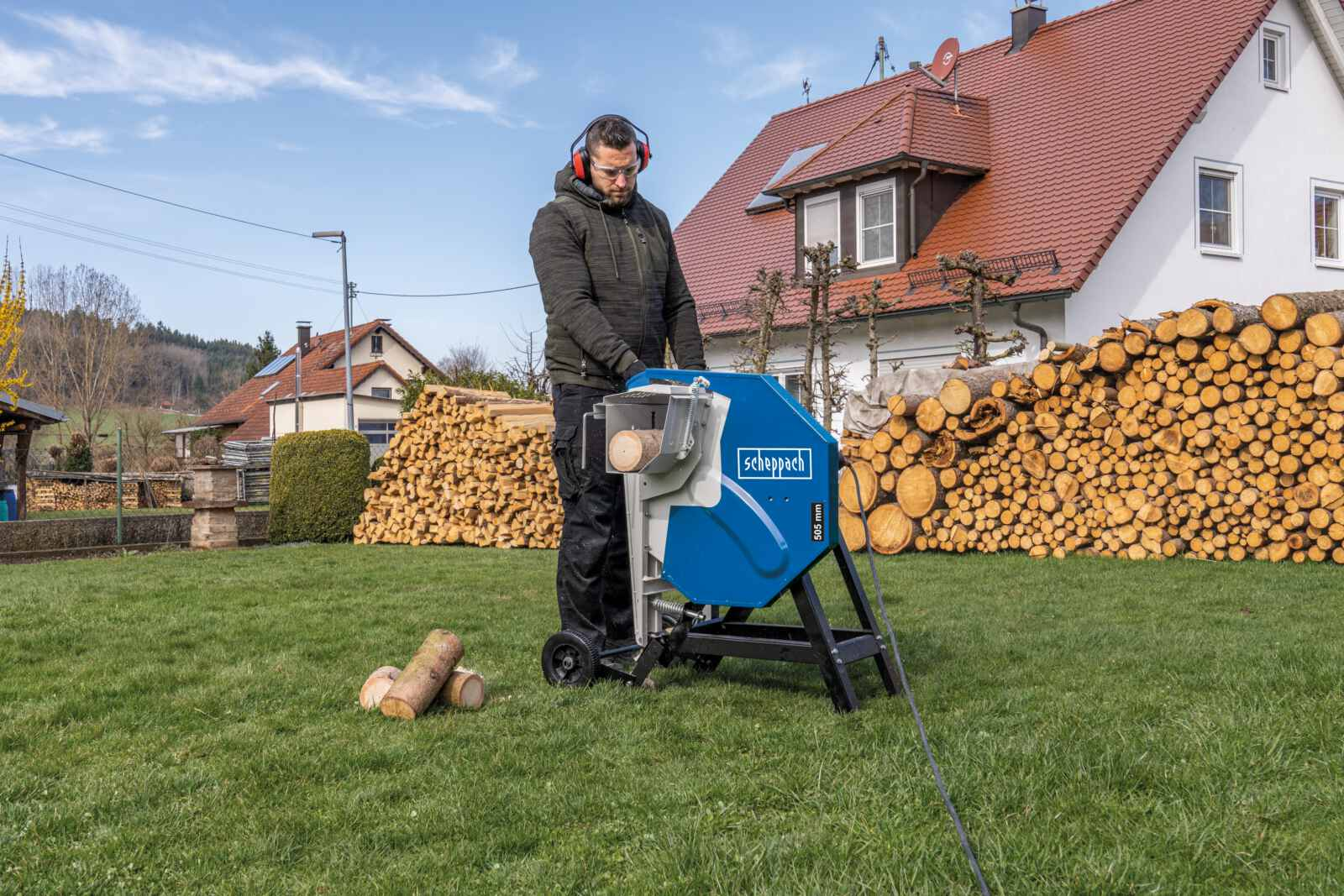 Mann benutzt blaue und schwarze Holzsäge im grasbewachsenen Garten. Holzscheite im Hintergrund.