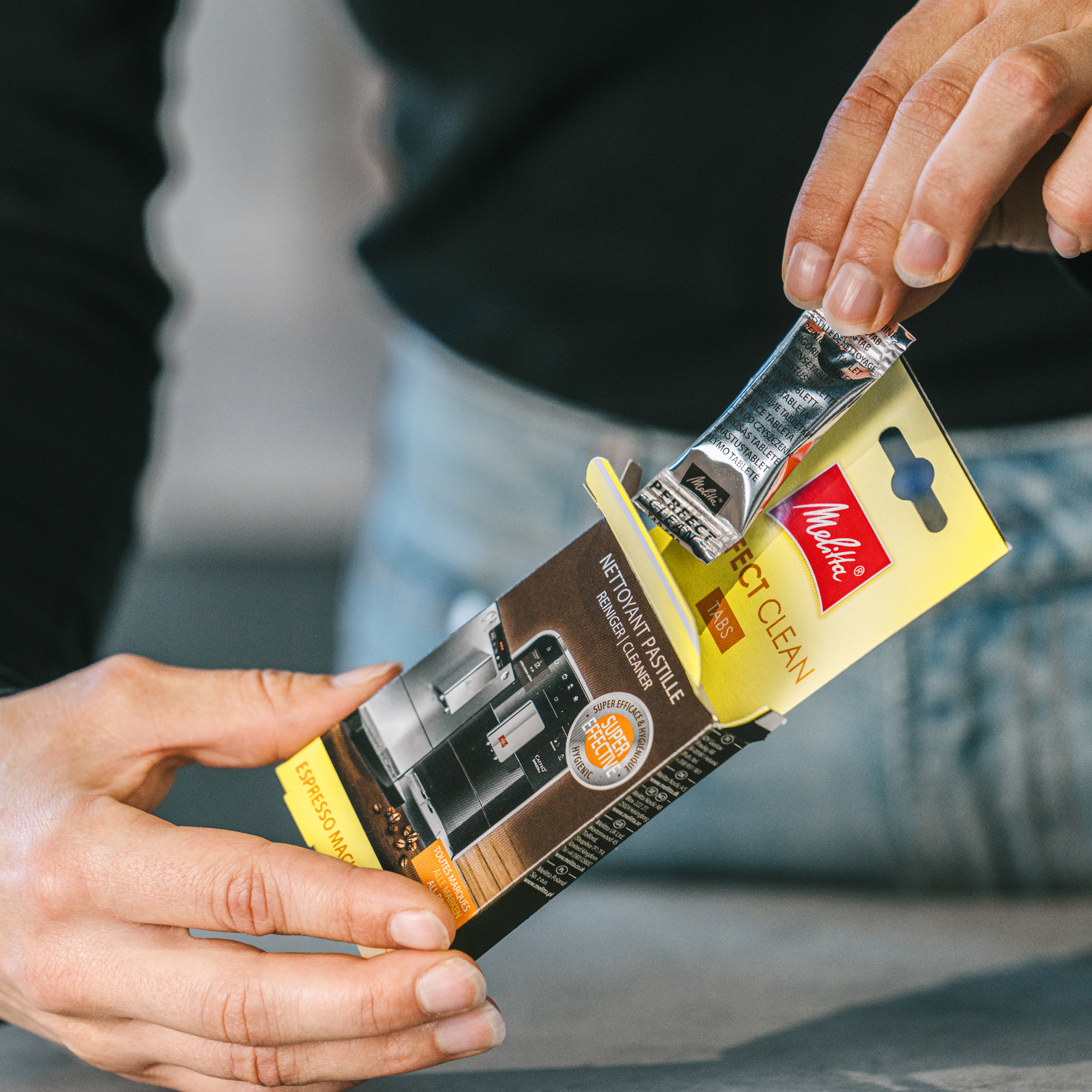 Manos de una persona abriendo un paquete de pastillas de limpieza Melitta Perfect Clean para máquina de espresso.