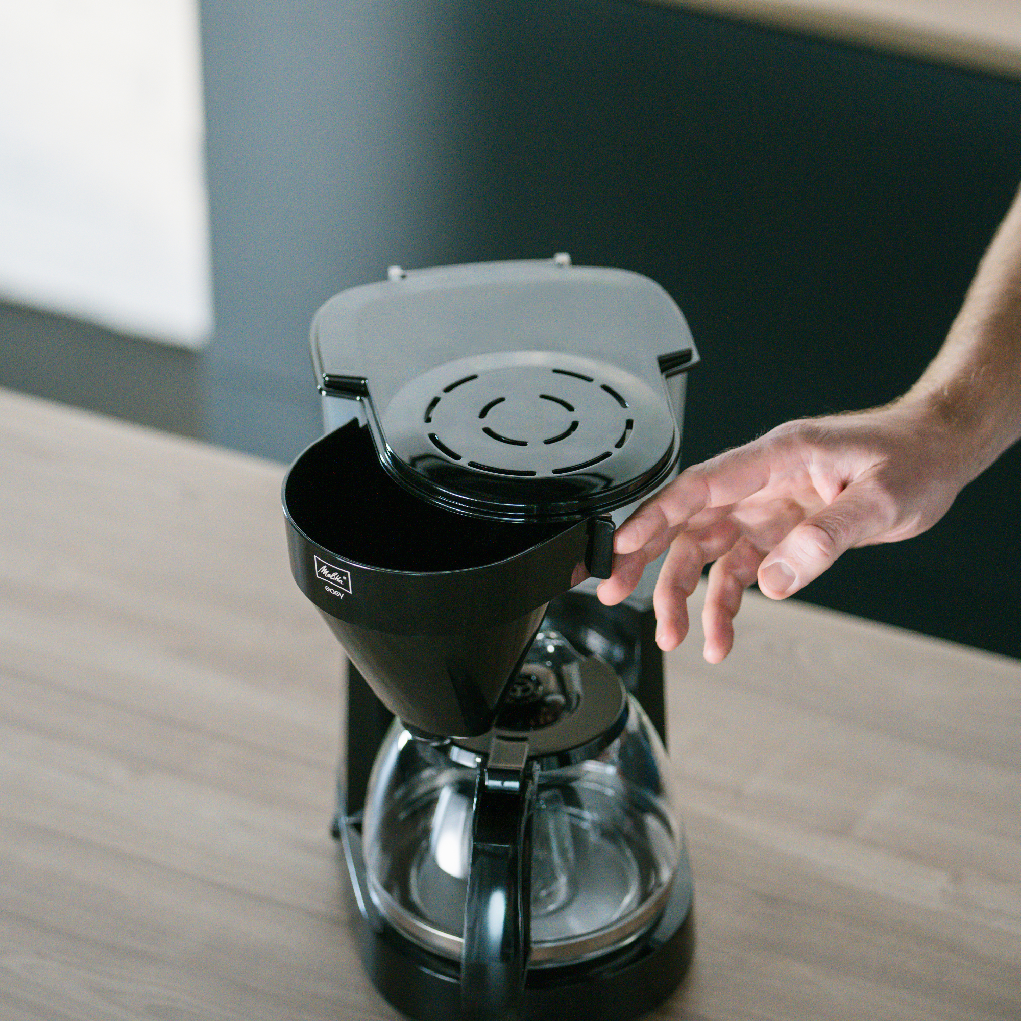 Ręka otwiera ekspres do kawy, aby umieścić kawę mieloną. Ekspres jest czarny i srebrny.