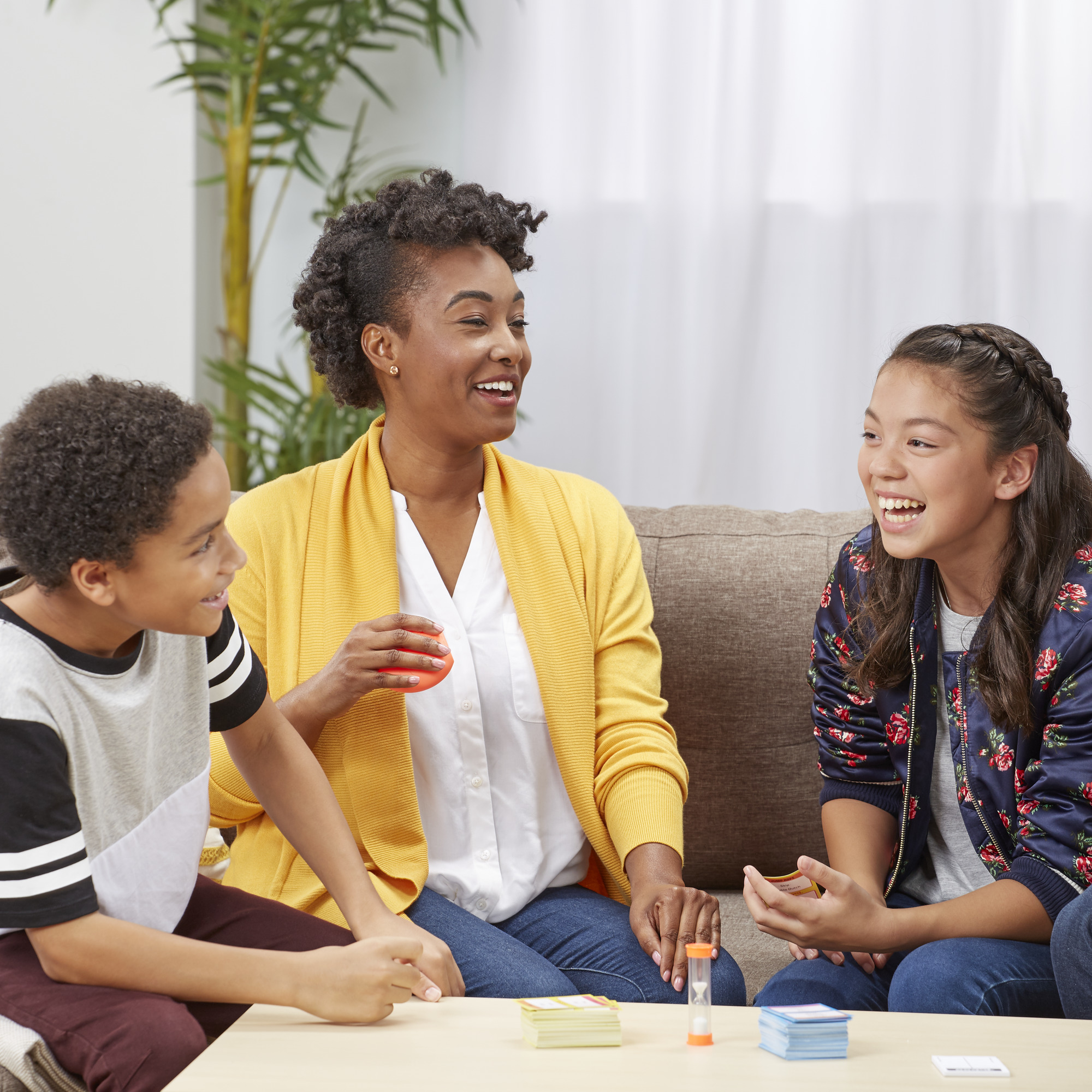 Drie personen, een vrouw en twee kinderen, lachen terwijl ze een spel spelen aan tafel. De vrouw houdt een rood voorwerp vast.