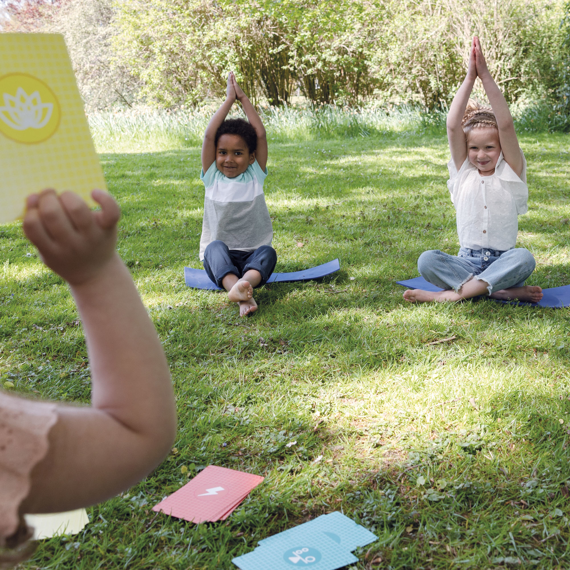 Bambini che fanno yoga sull'erba. Una donna tiene delle carte. Carte e alberi sullo sfondo.