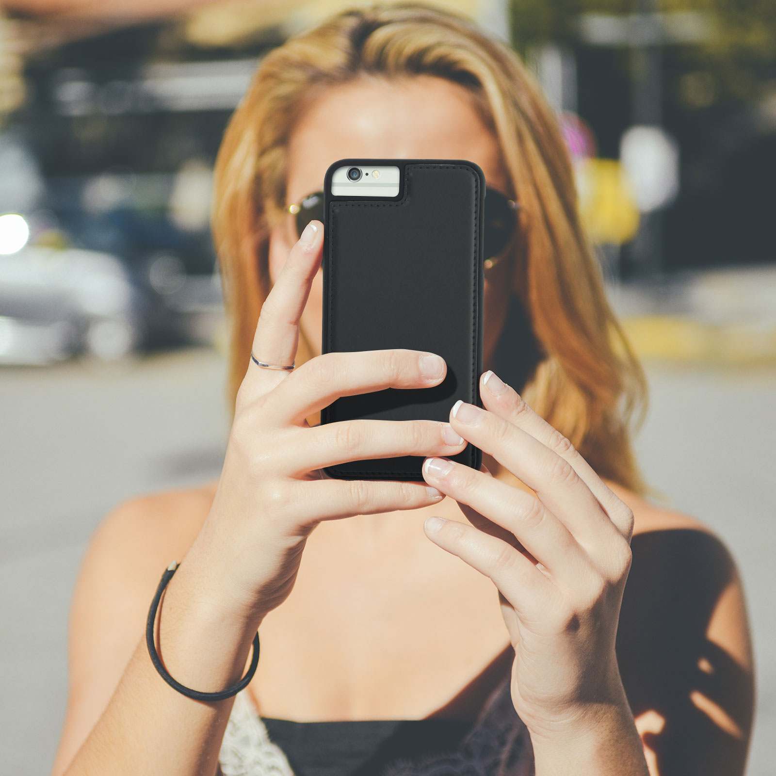 Donna che tiene un telefono nero, scattando una foto. Occhiali da sole, braccialetto e anello visibili.