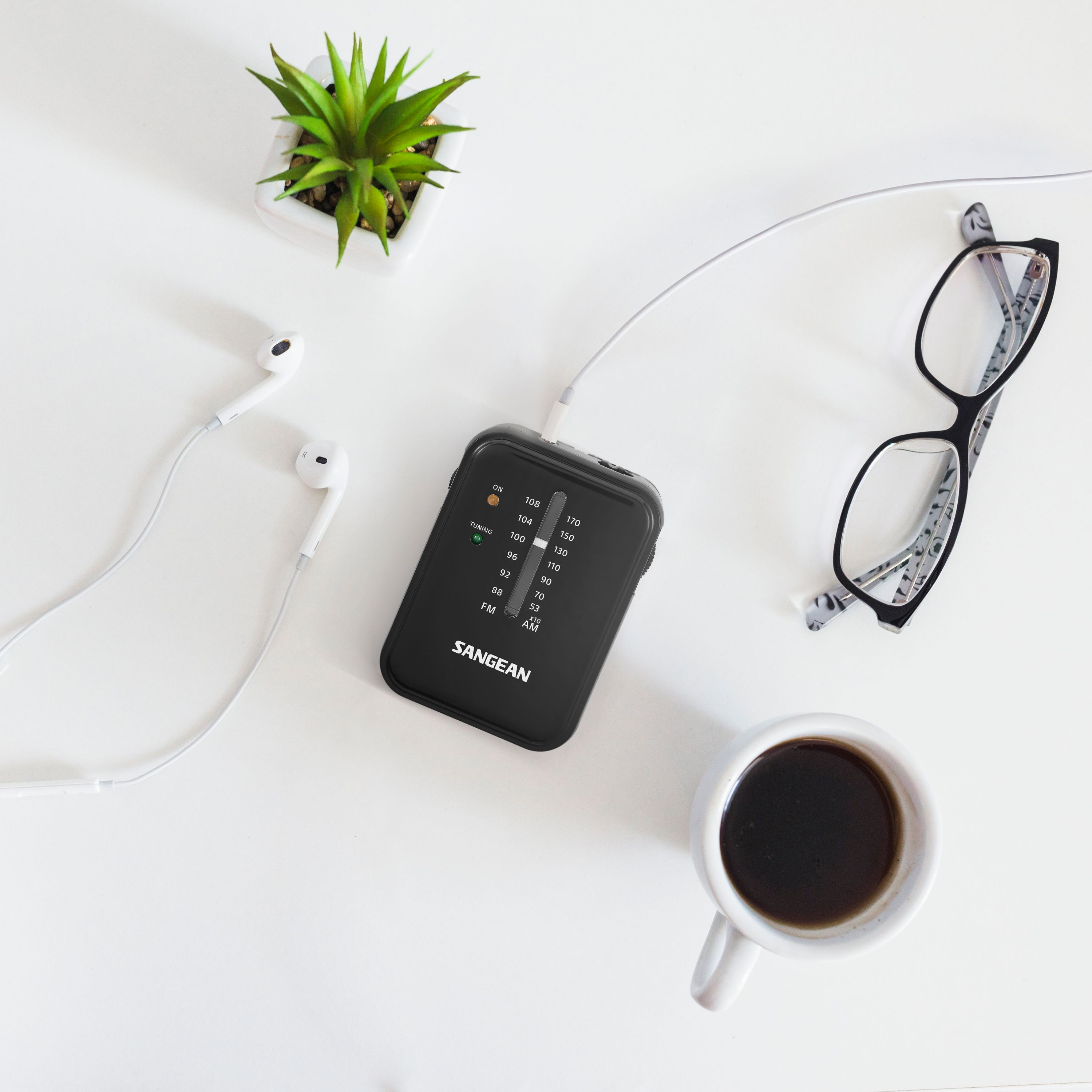 Czarne radio, słuchawki, okulary, kawa i roślina na białej powierzchni.