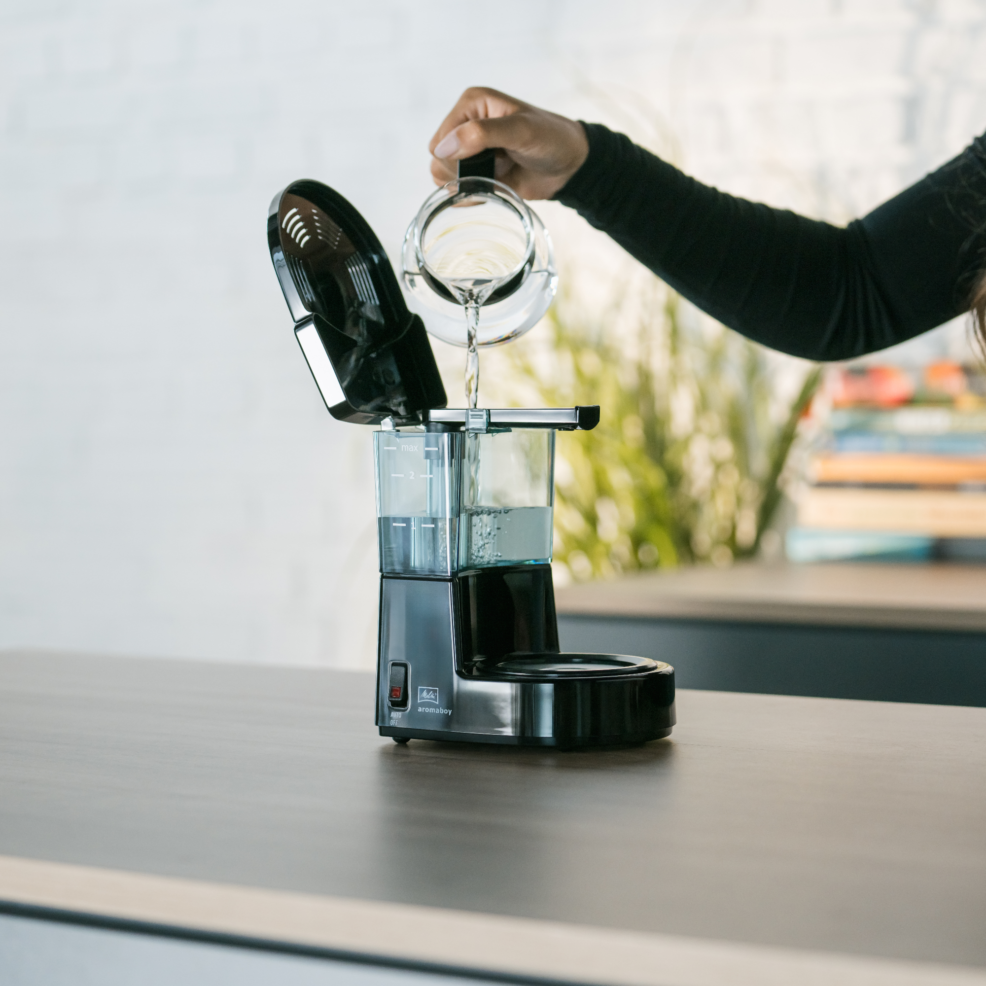 Osoba wlewa wodę do czarnego ekspresu do kawy, na drewnianym stole.