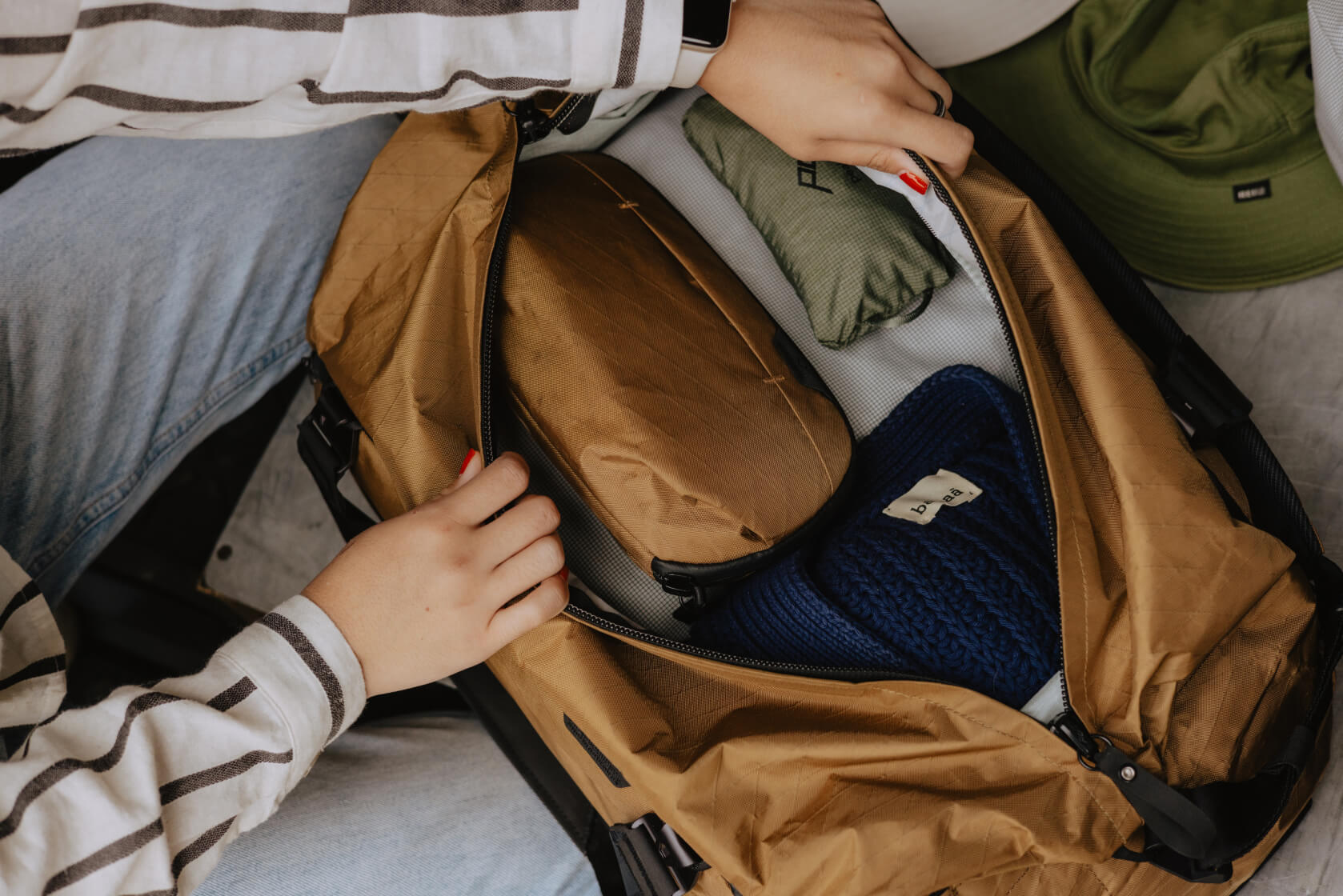 Osoba pakuje brązową torbę podróżną. Zawiera przedmioty, np. niebieską czapkę i brązową saszetkę.