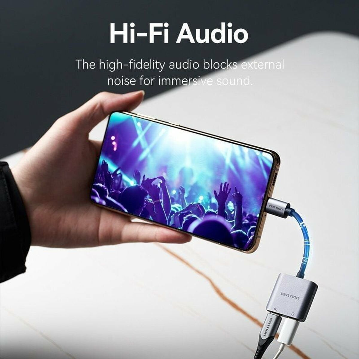 Una mano sosteniendo un teléfono con un adaptador de audio conectado. El teléfono muestra una multitud.
