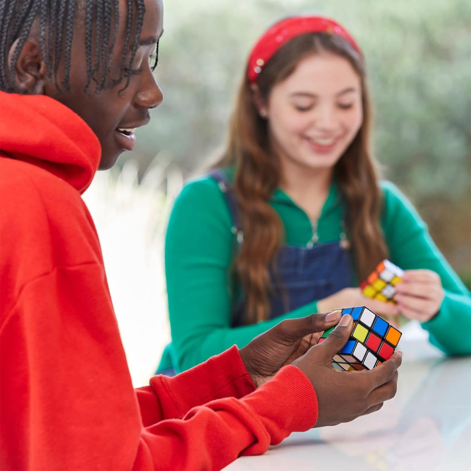 Zwei Personen, eine mit rotem Kapuzenpullover, lösen Rubik's Cubes, lächeln und konzentriert.