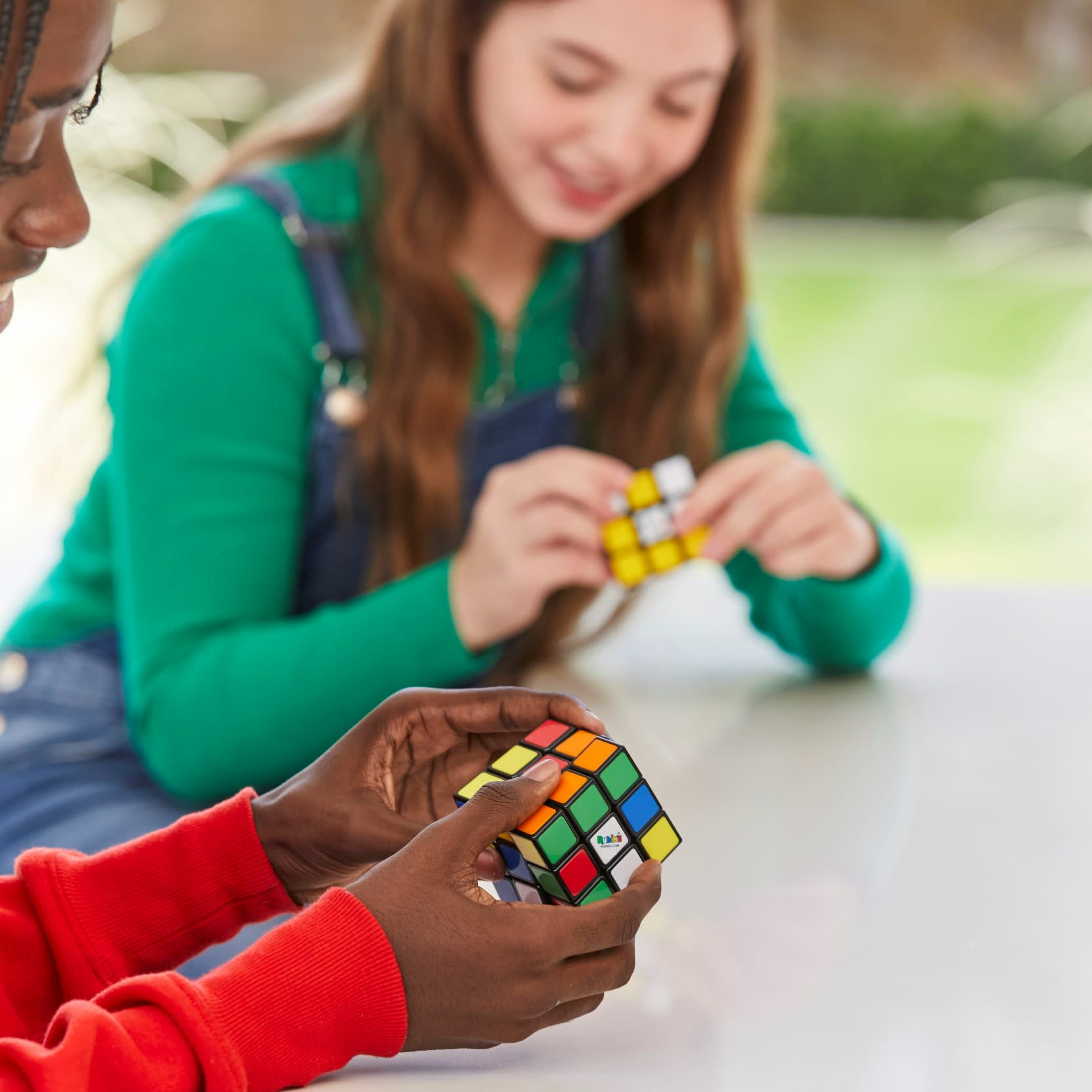 Zwei Personen lösen Rubik's Cubes. Eine hält einen Würfel in der Hand, die andere ist im Hintergrund verschwommen.