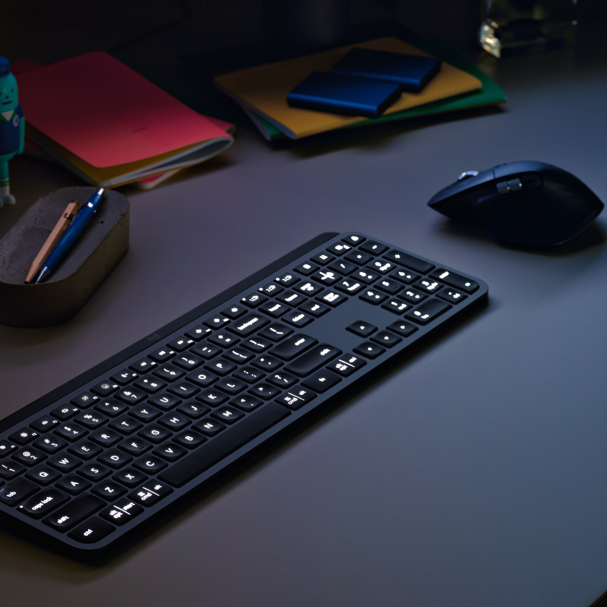 Tastiera nera illuminata su una scrivania scura. Sono visibili alcuni fogli, una penna e un mouse.