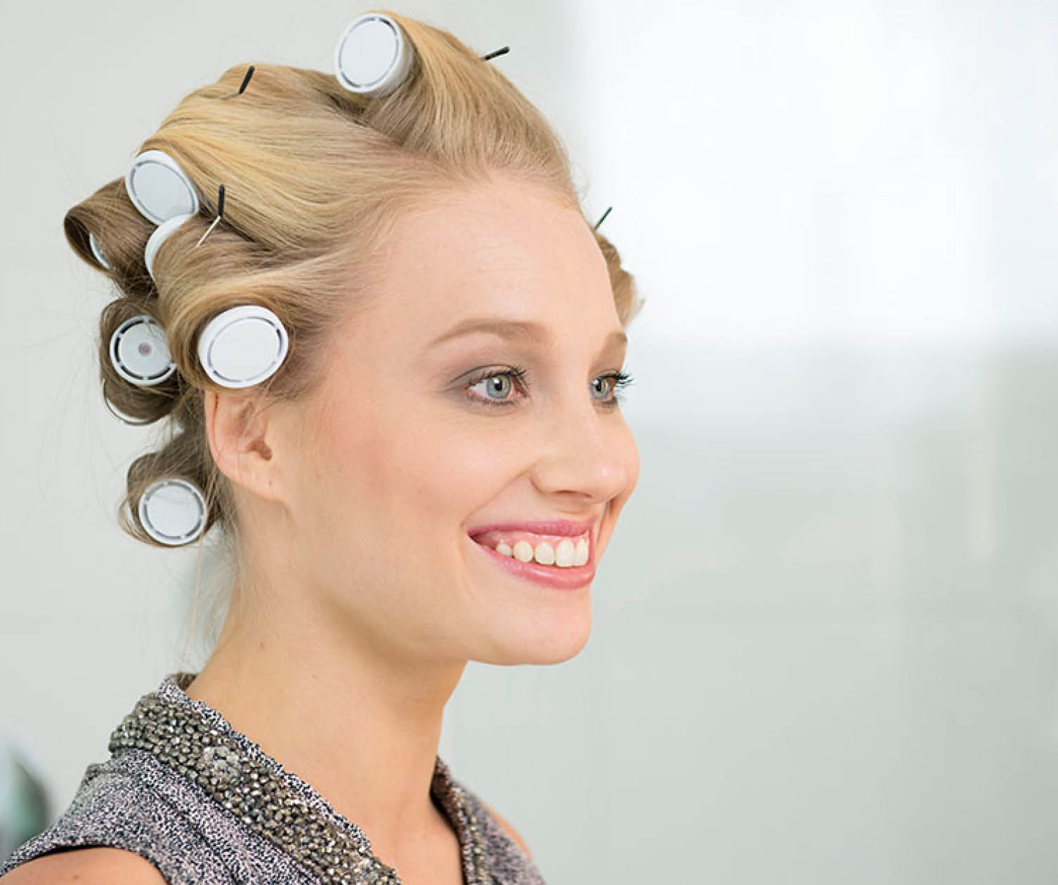 Blonde Frau mit Lockenwicklern lächelt. Hintergrund ist eine weiße Wand.