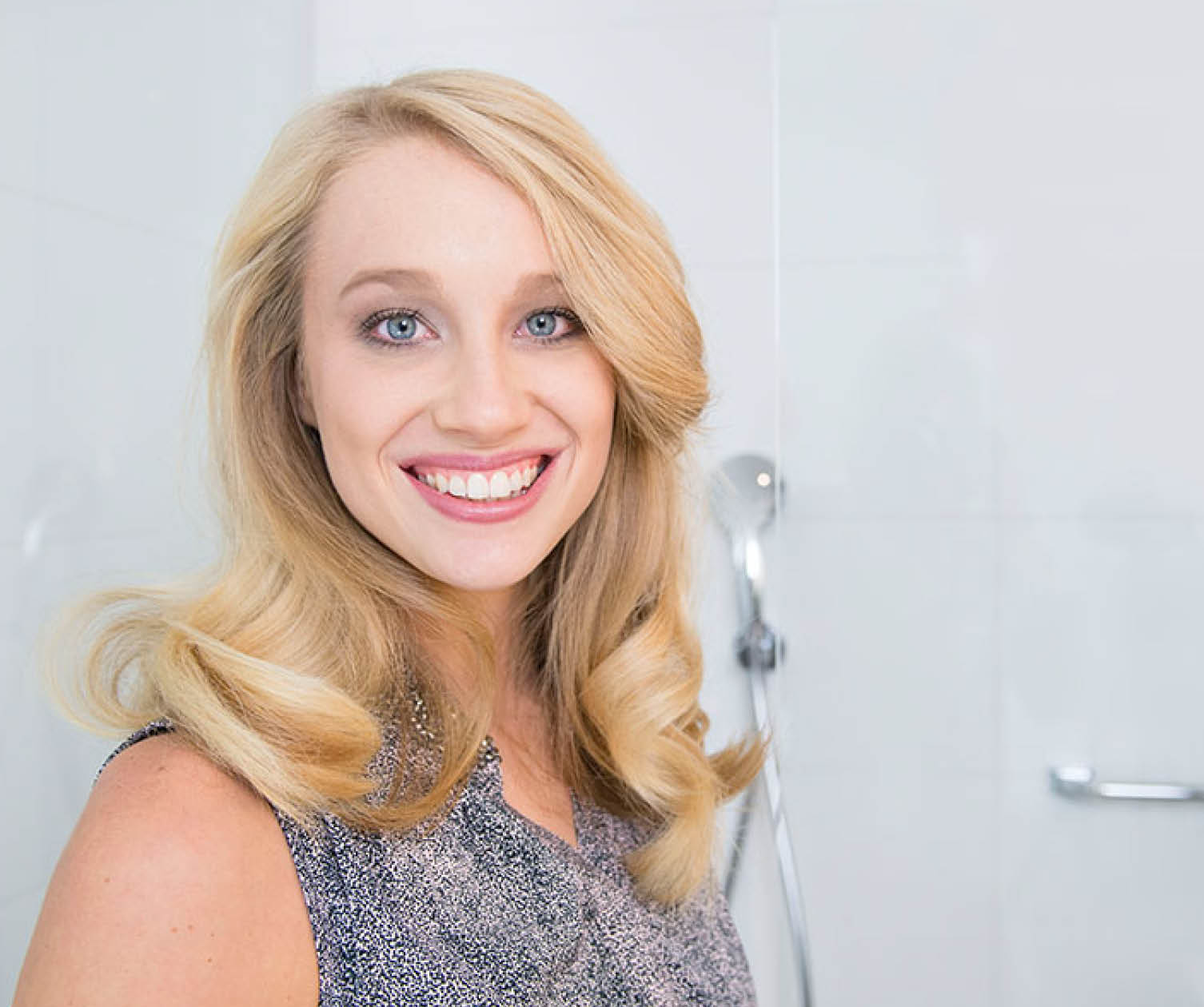 Lächelnde blonde Frau. Hintergrund ist ein weißes Badezimmer mit Duschkopf.