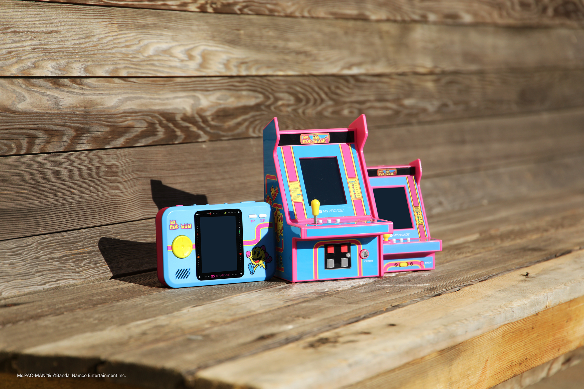 Trzy automaty do gier zręcznościowych retro na drewnianej powierzchni. Są niebieskie i różowe.