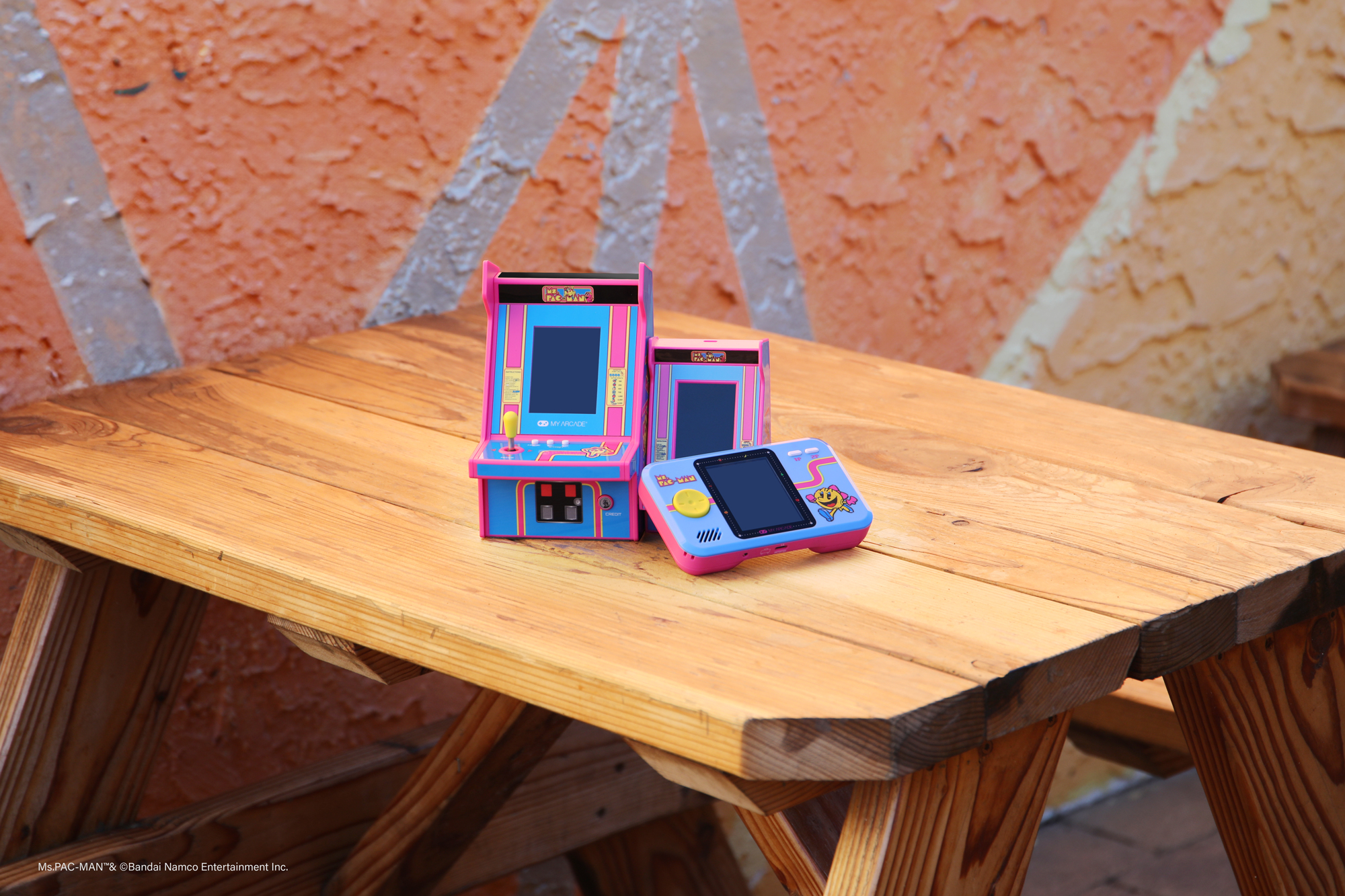 Trzy miniaturowe konsole do gier arkadowych i kontroler na drewnianym stole. Tłem jest pomarańczowa ściana.