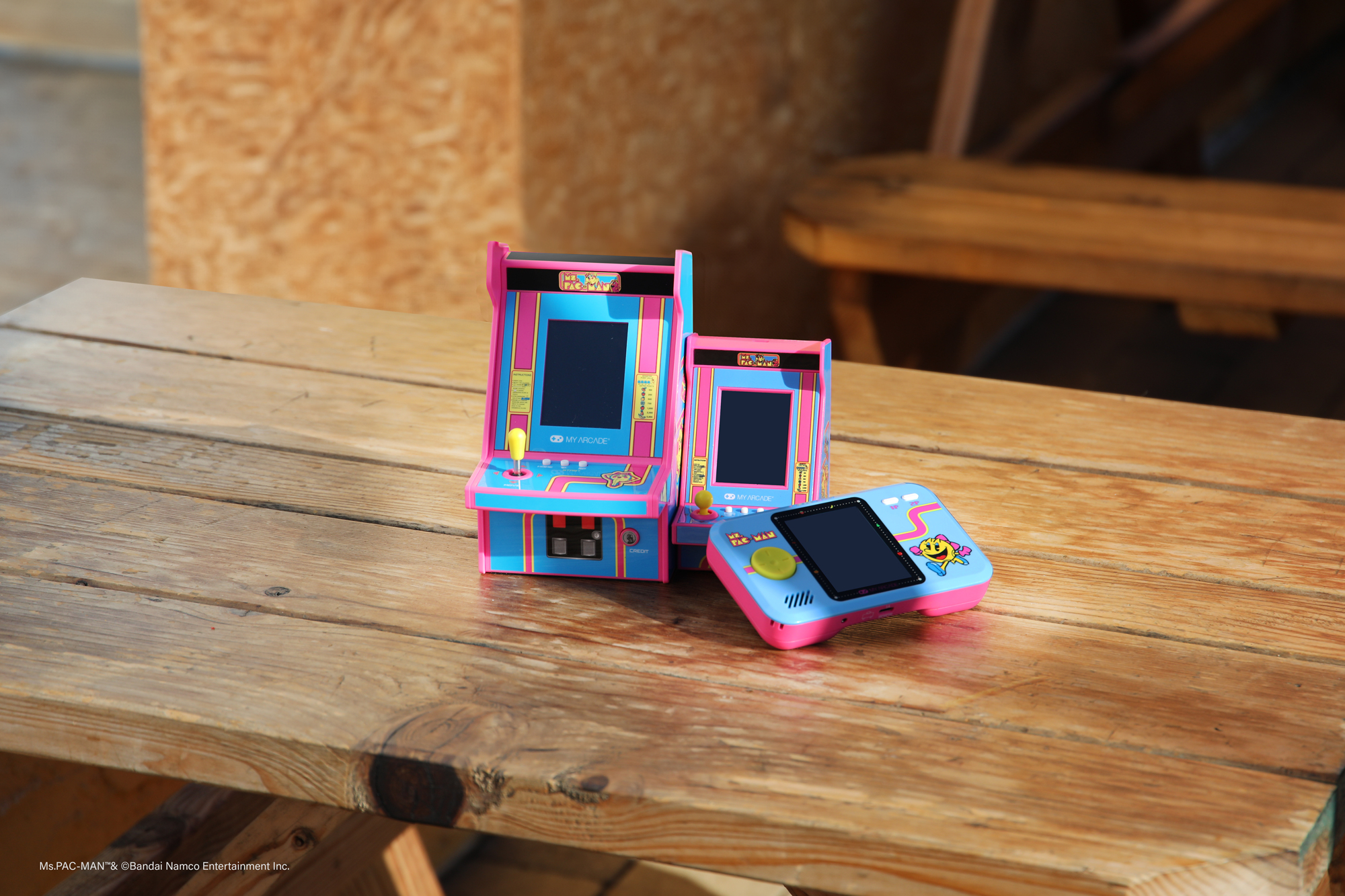 Trzy miniaturowe konsole do gier arkadowych i kontroler na drewnianym stole. Tło to drewniana konstrukcja.