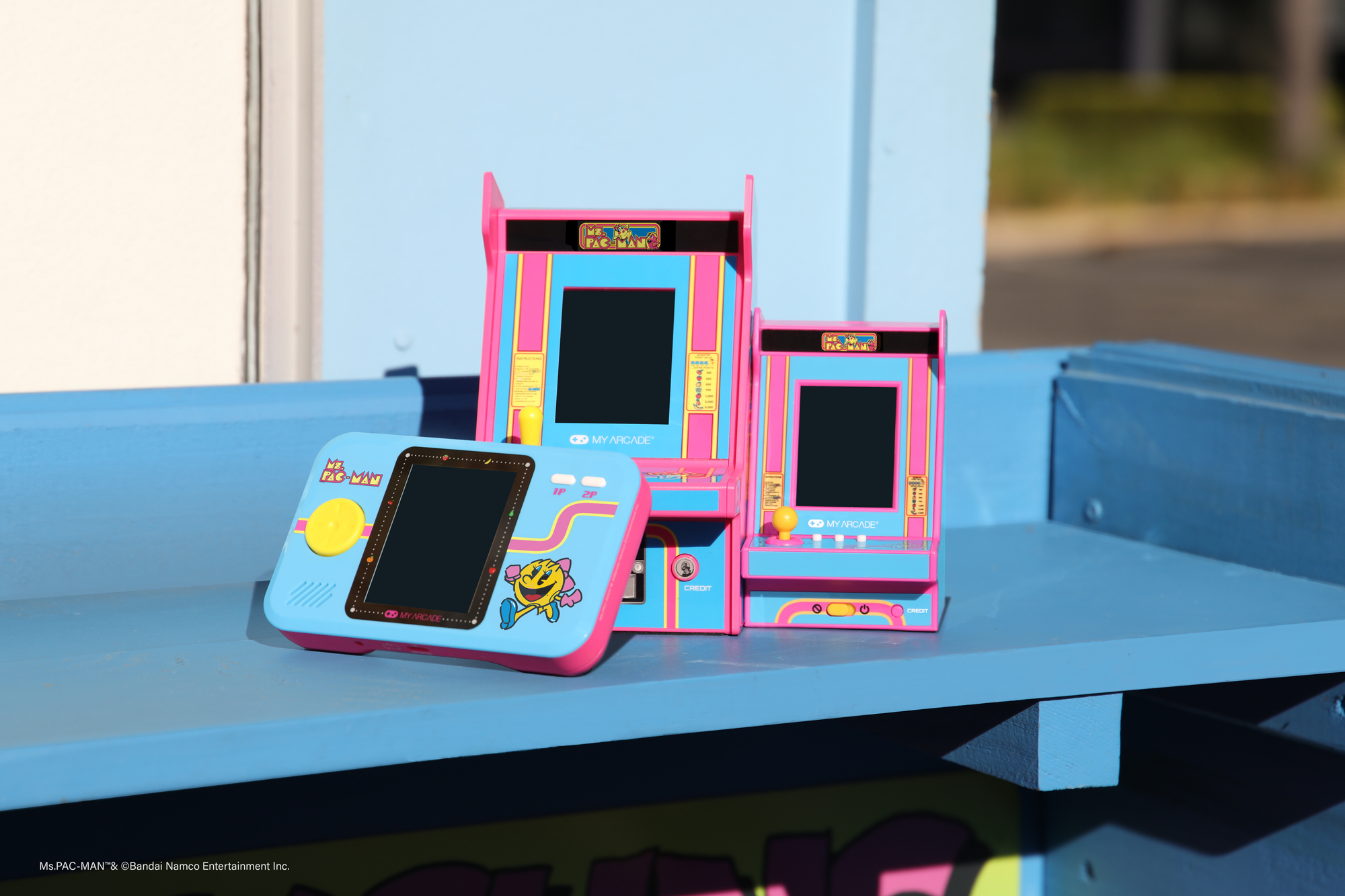 Trzy kolorowe konsole do gier Ms. Pac-Man na niebieskiej półce. Jedna przenośna, dwie wersje stołowe, wszystkie w różu i niebieskim.