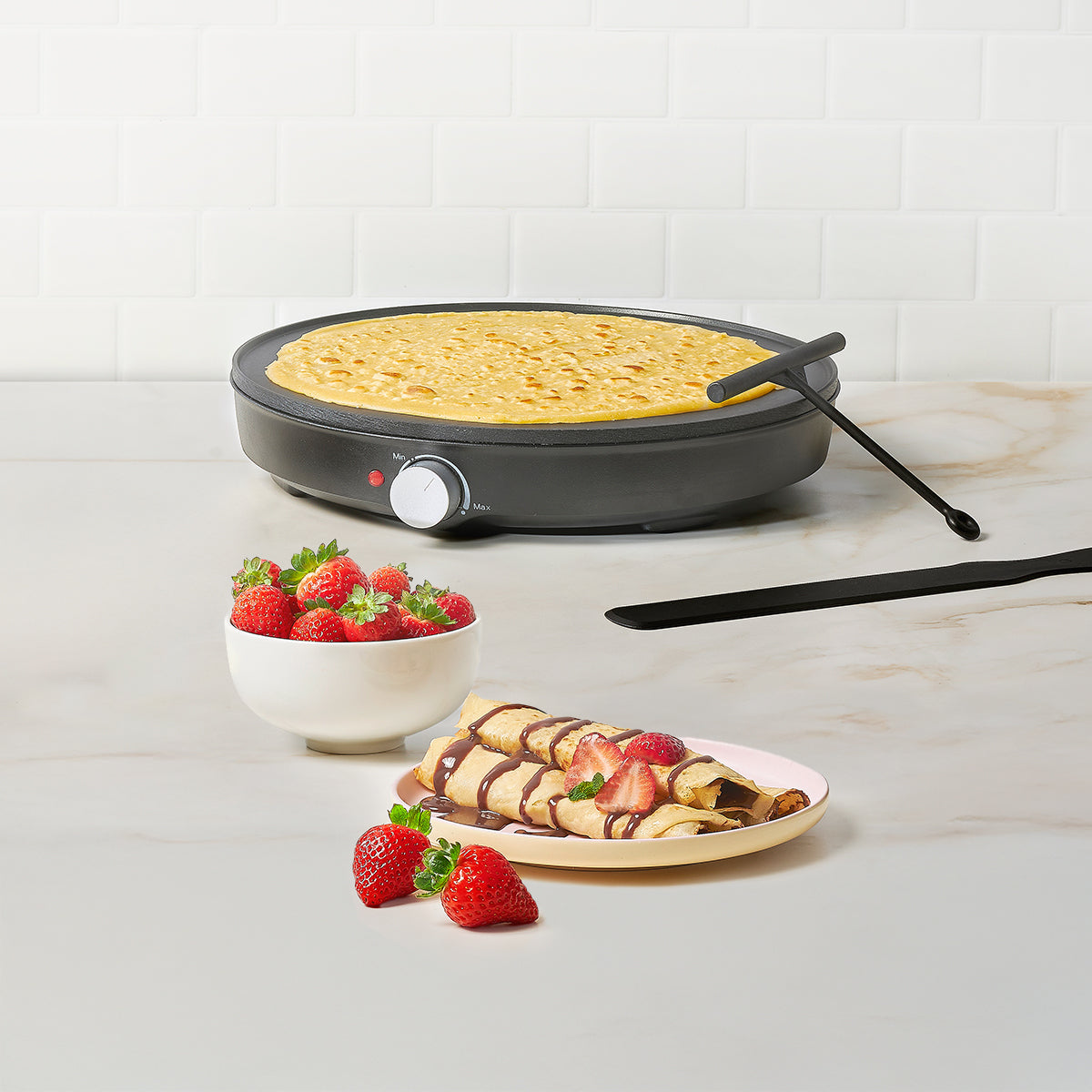 Crepera con crepes, fresas y chocolate. Escena de cocina, fondo de azulejos blancos.