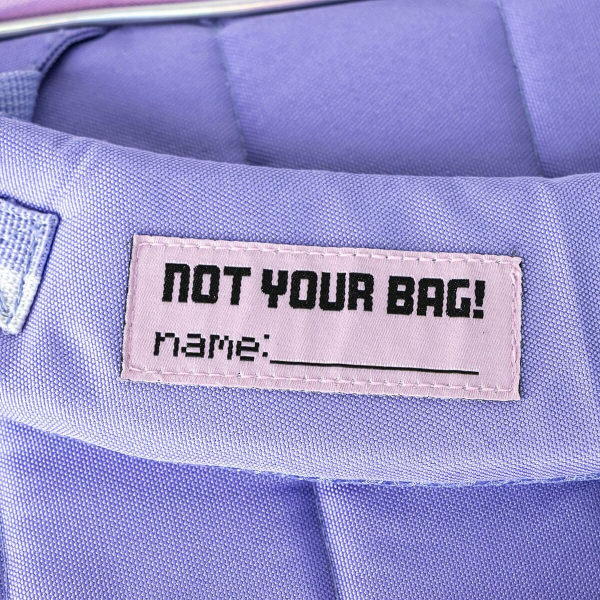 Bolso lavanda con una etiqueta que dice 'NOT YOUR BAG!' y sección de nombre.