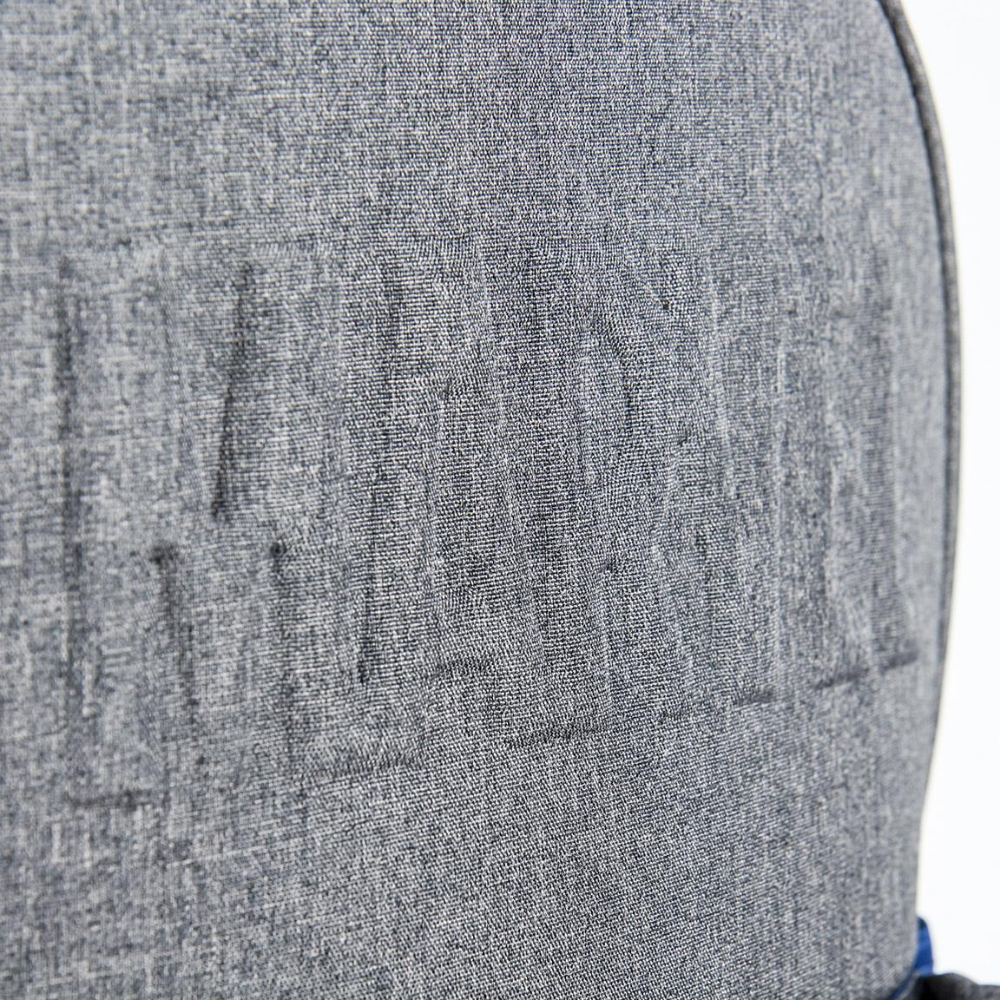 Primer plano de una tela gris, mostrando parcialmente una mochila. Presenta un diseño en relieve sutil.