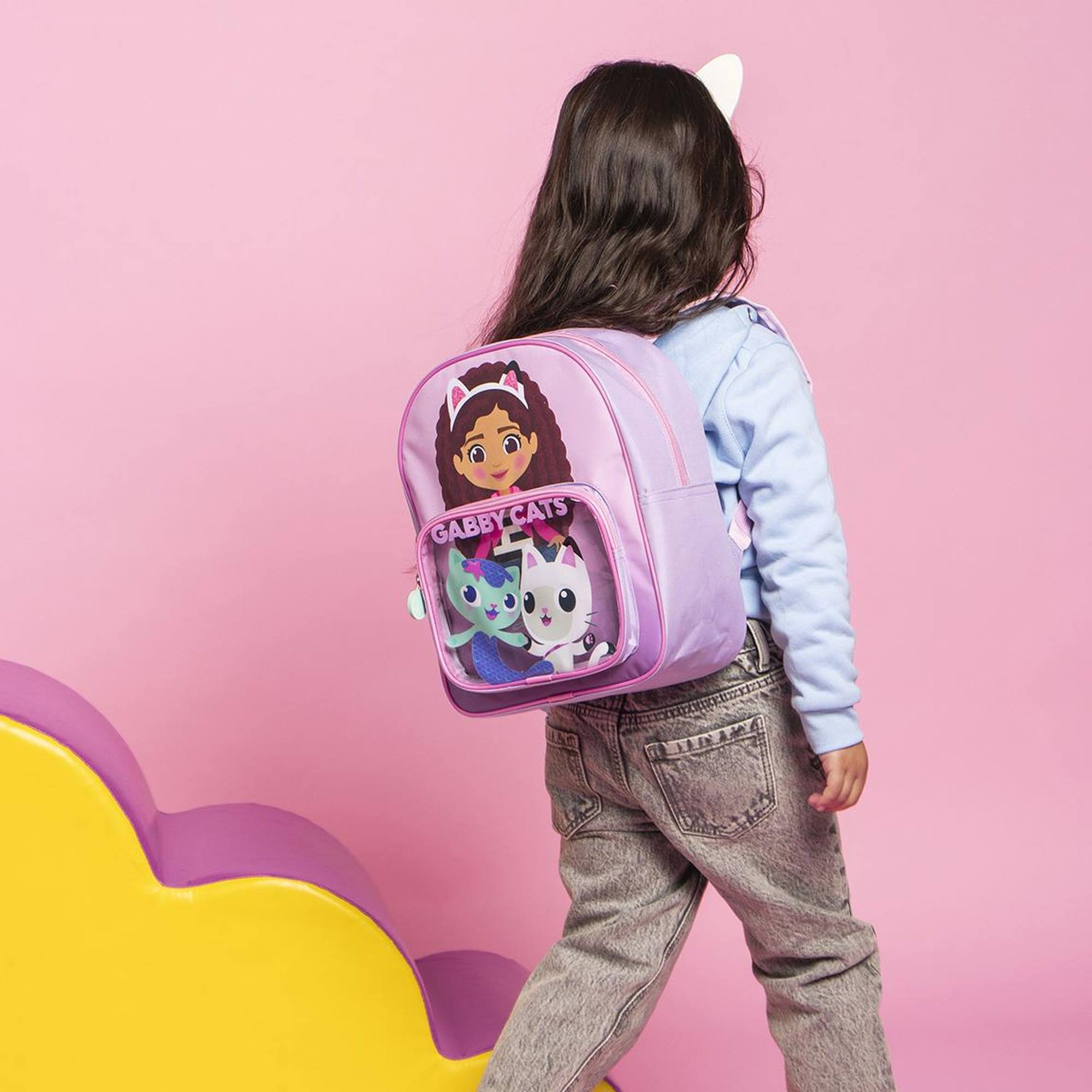 Una niña lleva una mochila de Gabby's Dollhouse. La mochila es morada y tiene a Gabby y Gabby Cats.