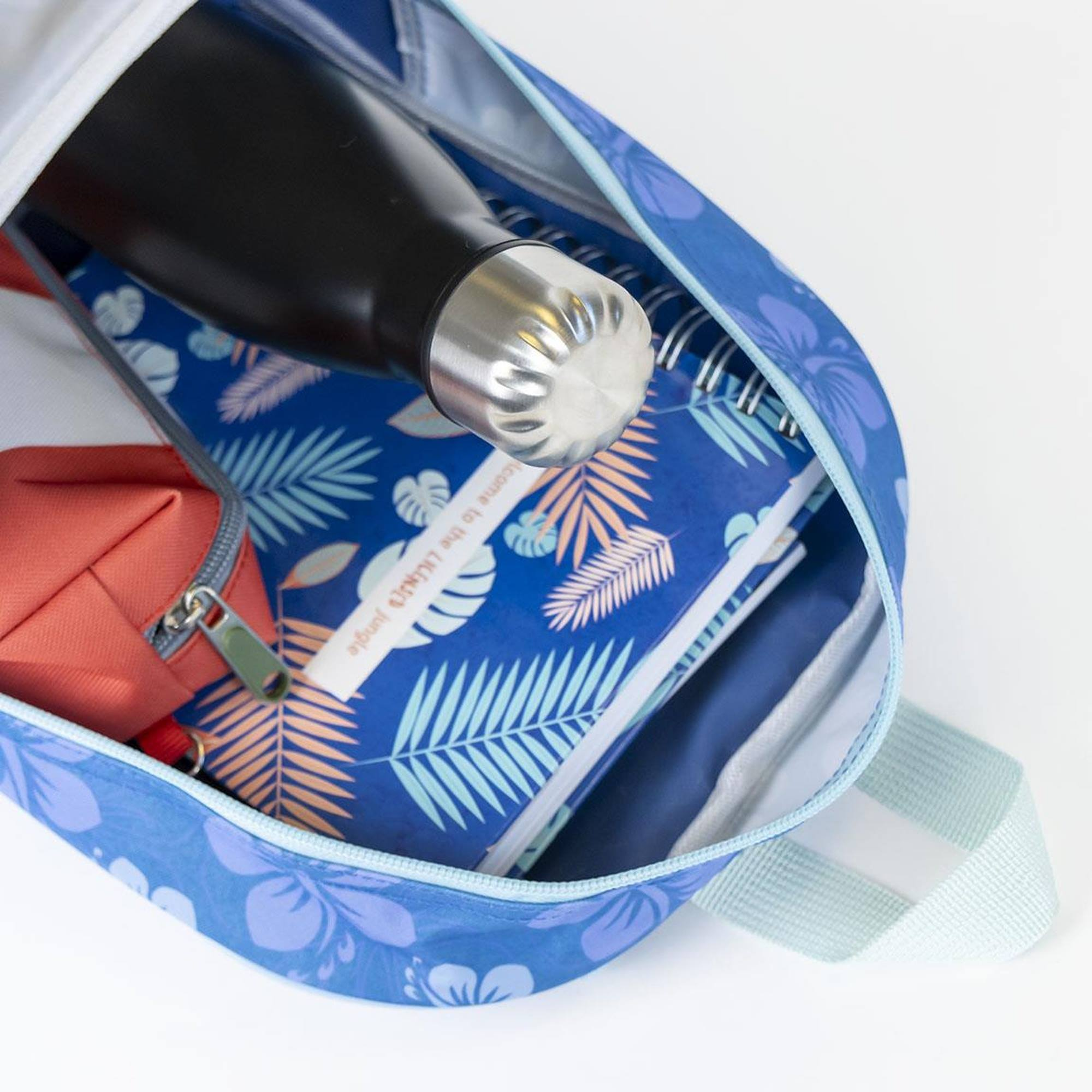 Offener blauer Rucksack, darin eine Wasserflasche, ein Federmäppchen und ein Notizbuch. Blaues Blumenmuster. Weißer Hintergrund.
