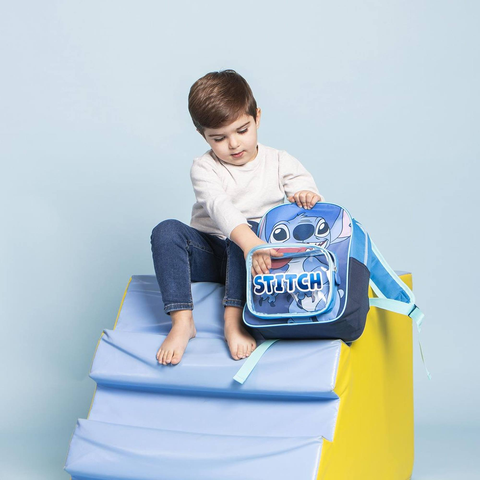 Un niño pequeño sentado en un escalón azul sosteniendo una mochila azul de Stitch. Fondo azul y amarillo.