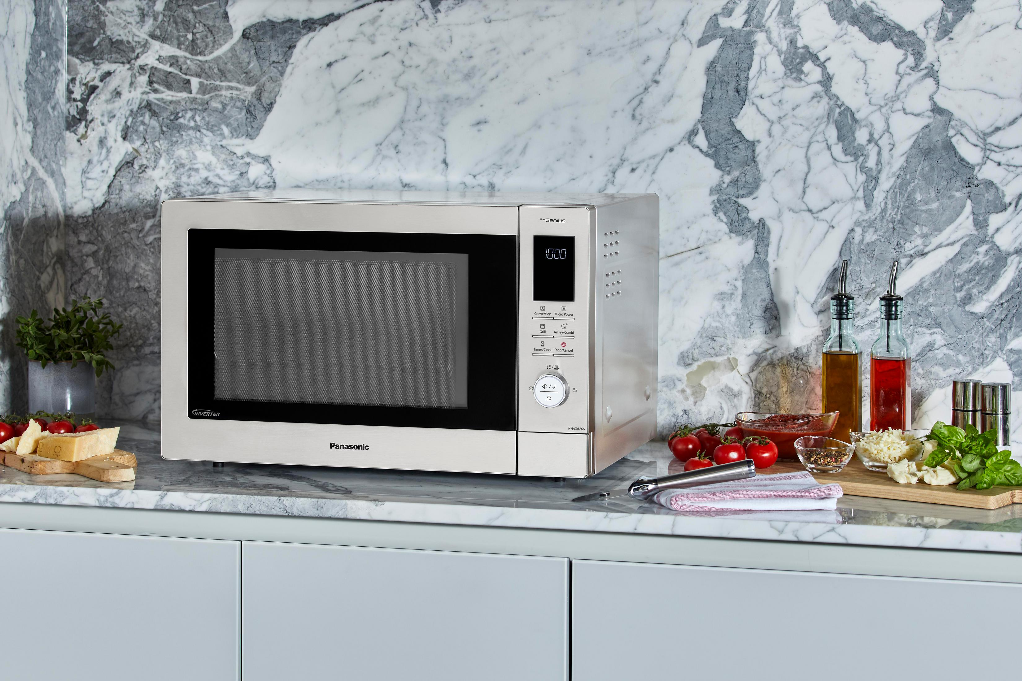 Kuchenka mikrofalowa Panasonic na marmurowym blacie z pomidorami, serem i butelkami oliwy z oliwek. Widoczne białe szafki.
