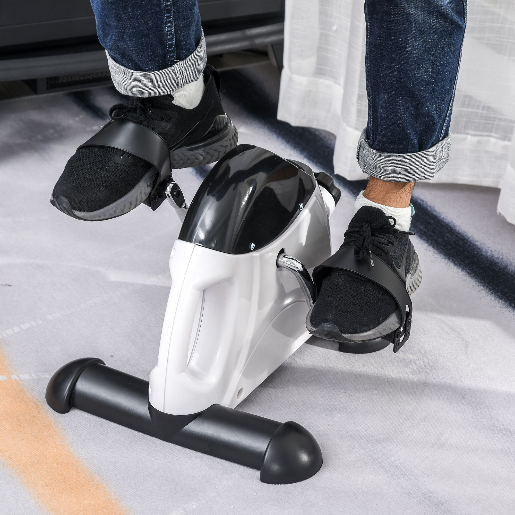 Person benutzt einen Mini-Heimtrainer. Beine treten, schwarze Schuhe. Das Fahrrad ist weiß und schwarz.