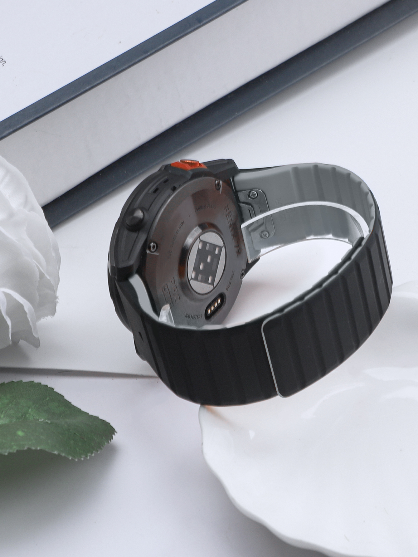 Rückseite einer schwarzen Smartwatch mit schwarzem Armband und orangefarbenem Knopf. Weißer Hintergrund.