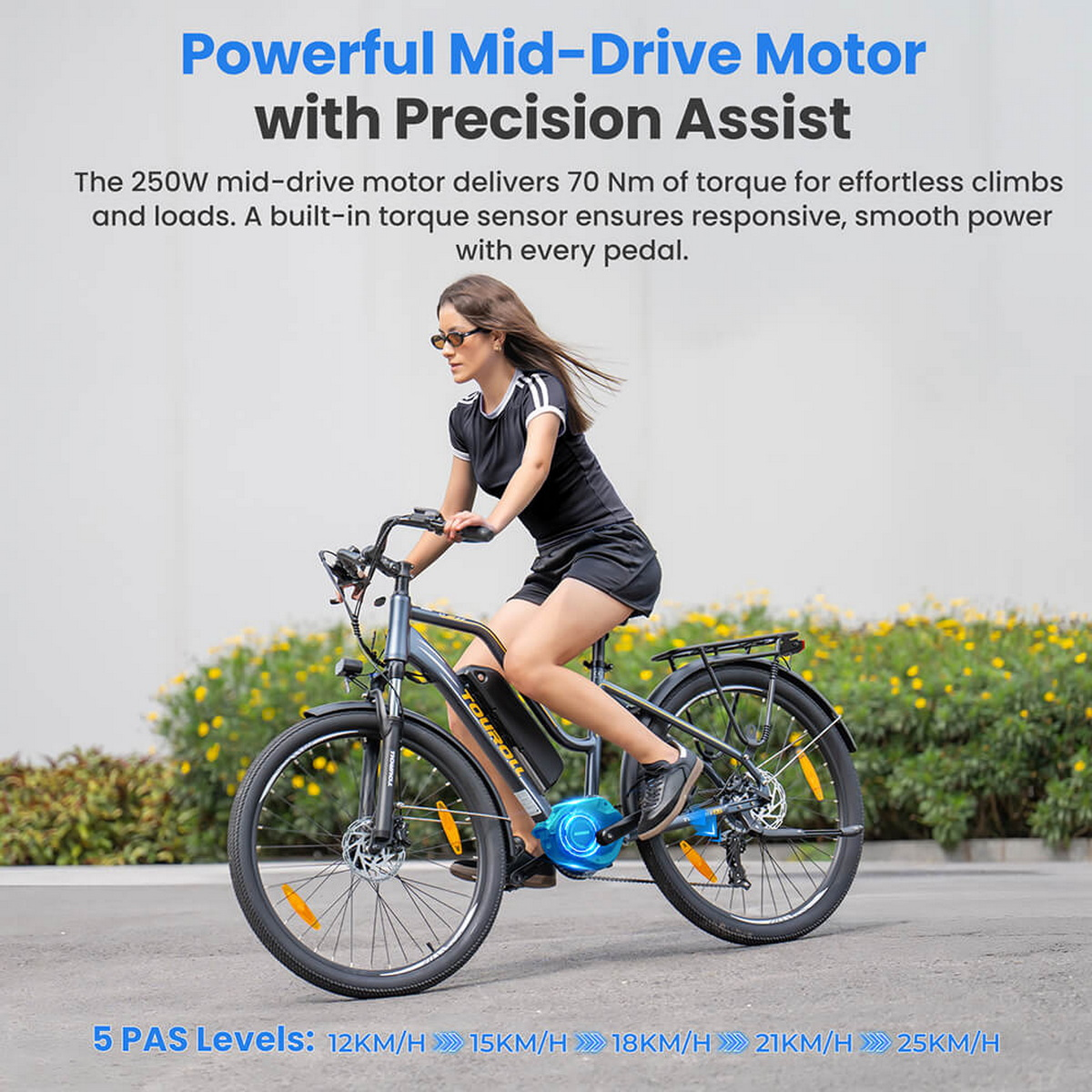 Kobieta jadąca rowerem elektrycznym po drodze. Rower jest szary i ma niebieski silnik. Po bokach są kwiaty.