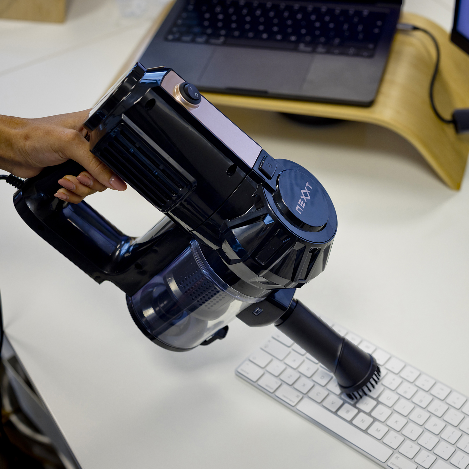 A black vacuum is held over a white keyboard. A laptop is on a wooden stand in the background.