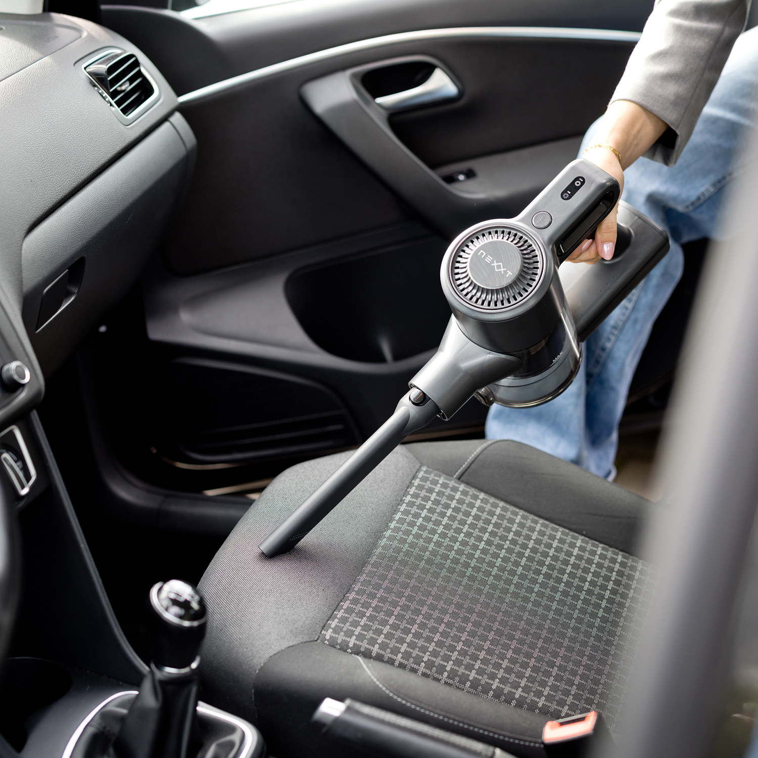A person vacuums a car's interior with a cordless vacuum cleaner, focusing on the seat.