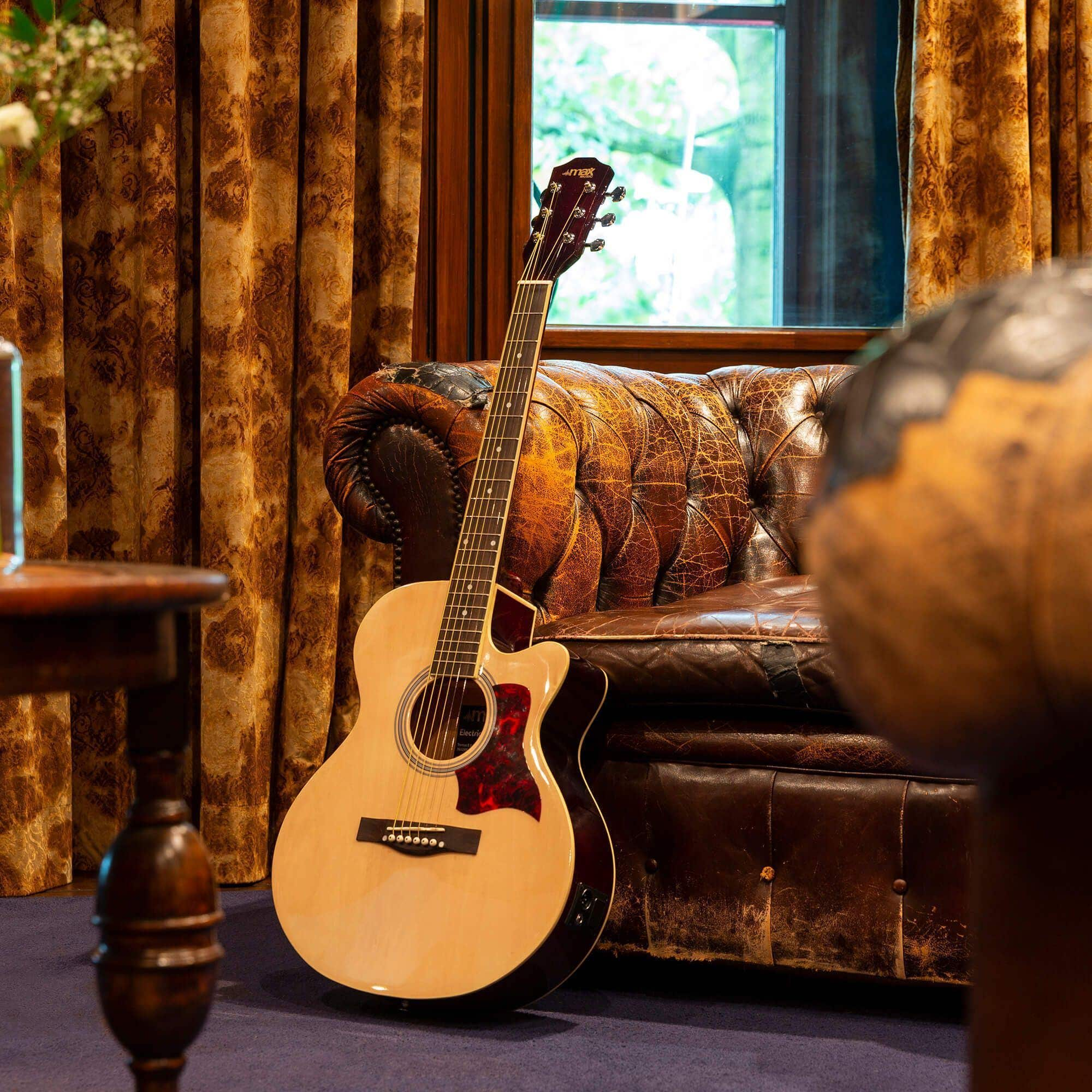 Eine Gitarre lehnt an einem braunen Ledersofa. Fenster und Vorhänge im Hintergrund sichtbar.