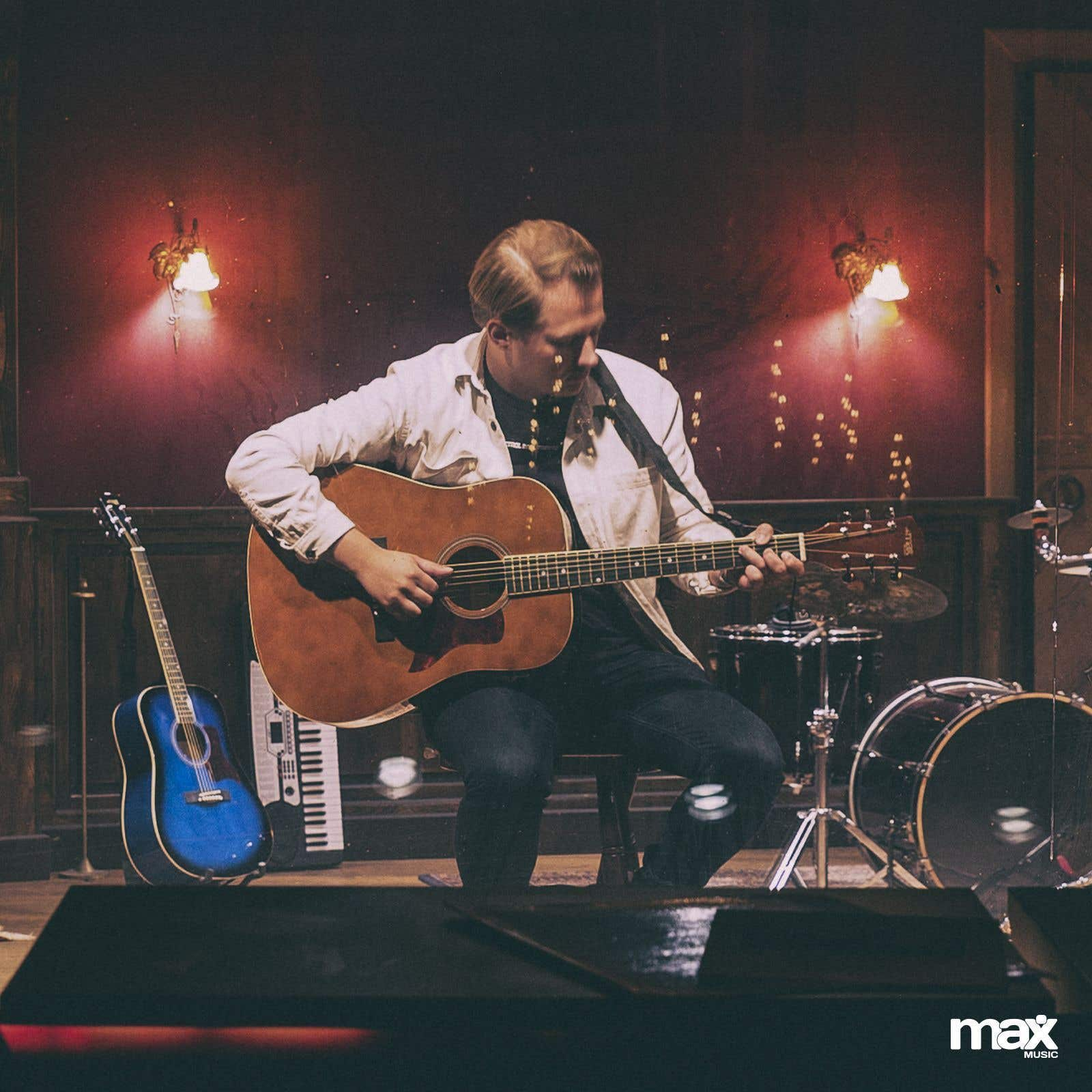 Man speelt akoestische gitaar. Podium, rode muur, blauwe gitaar, keyboard, drums. Gedimde verlichting.